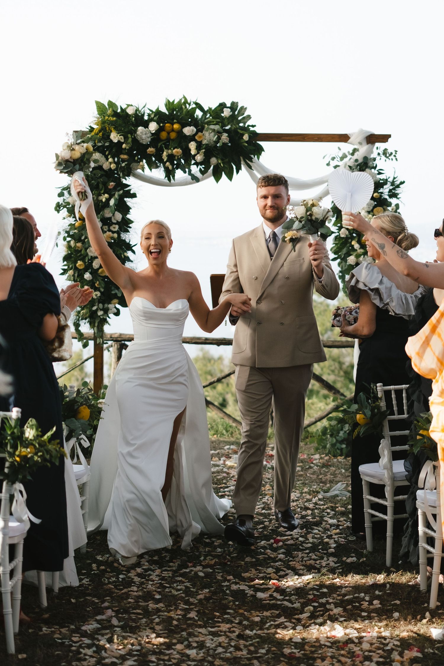 Joyful newlyweds celebrate recessional under floral arch with raised hands at outdoor wedding ceremony.