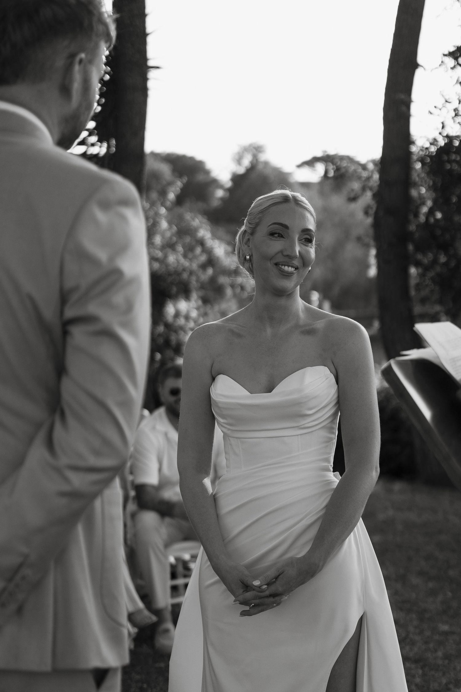Smiling bride in strapless white gown stands at outdoor wedding ceremony facing her groom.