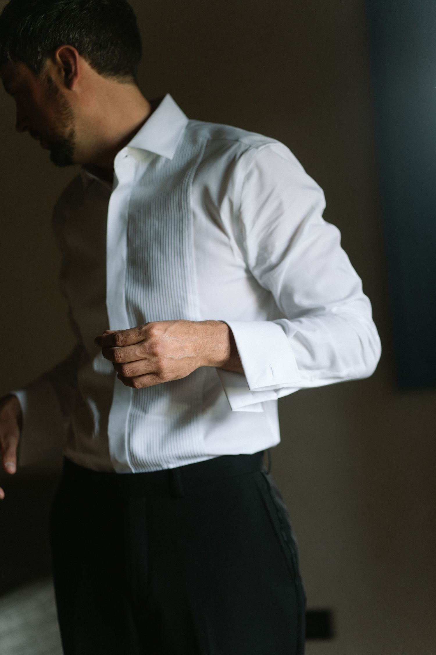 Groom in white tuxedo shirt with French cuffs fastening his cufflinks while getting ready for wedding.