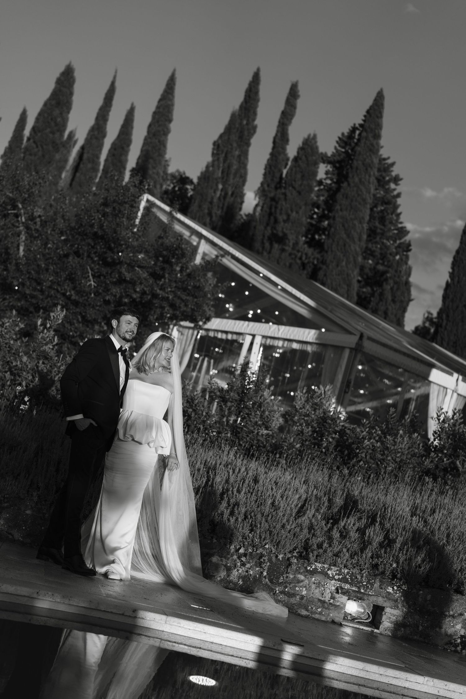 Black and white wedding photo of bride and groom standing near a pool with tall cypress trees at dusk.