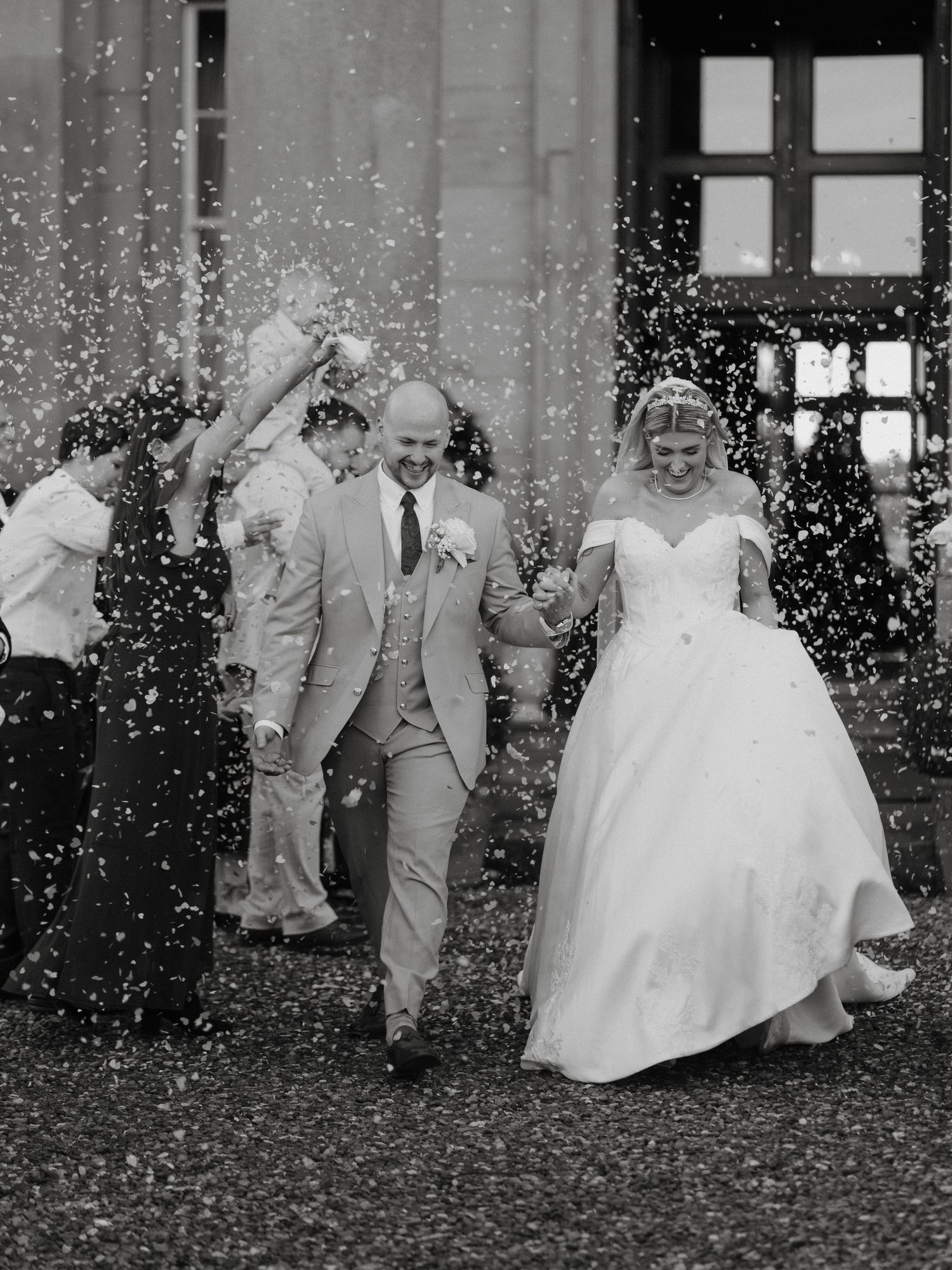 Black and white photo of a bride and groom walking through confetti shower outside a grand building.