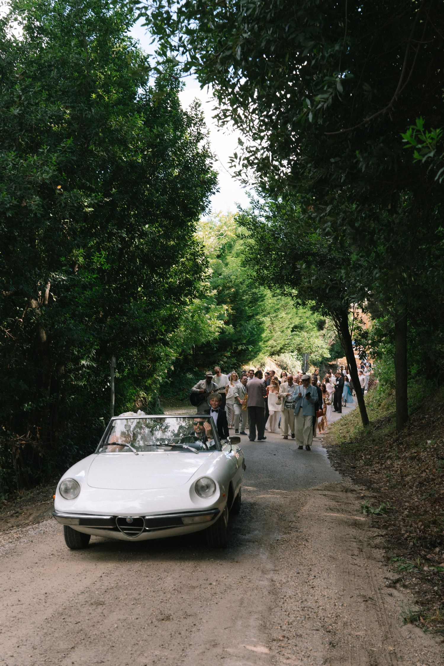 Wedding guests follow a classic white Alfa Romeo Spider convertible down a tree-lined dirt road.
