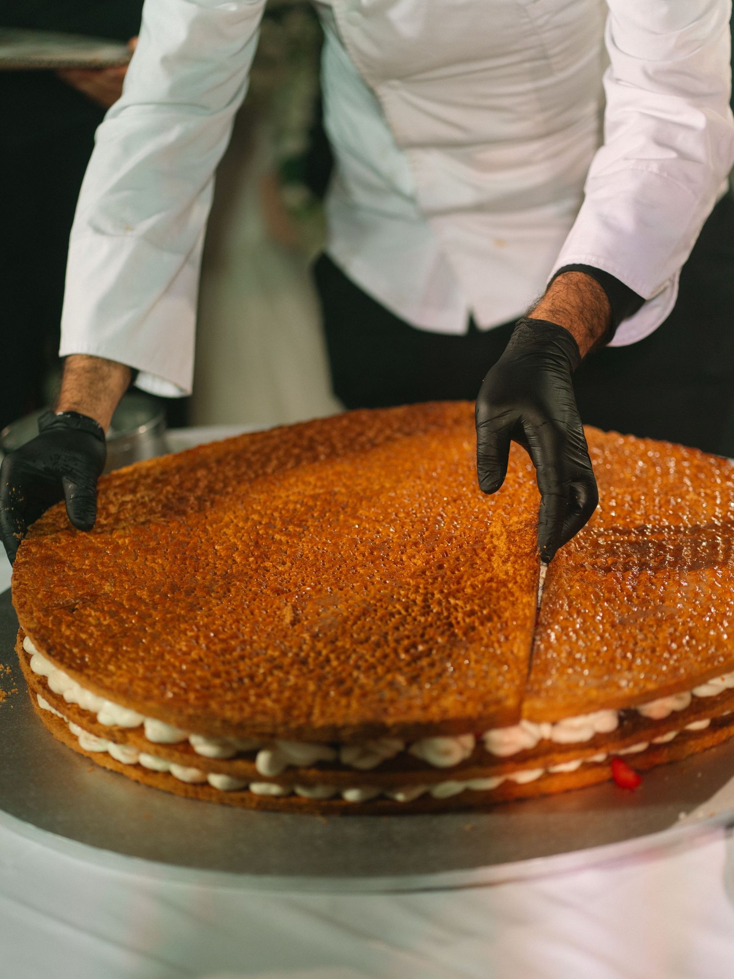Chef in black gloves cutting a large layered honey cake with a knife at a wedding.
