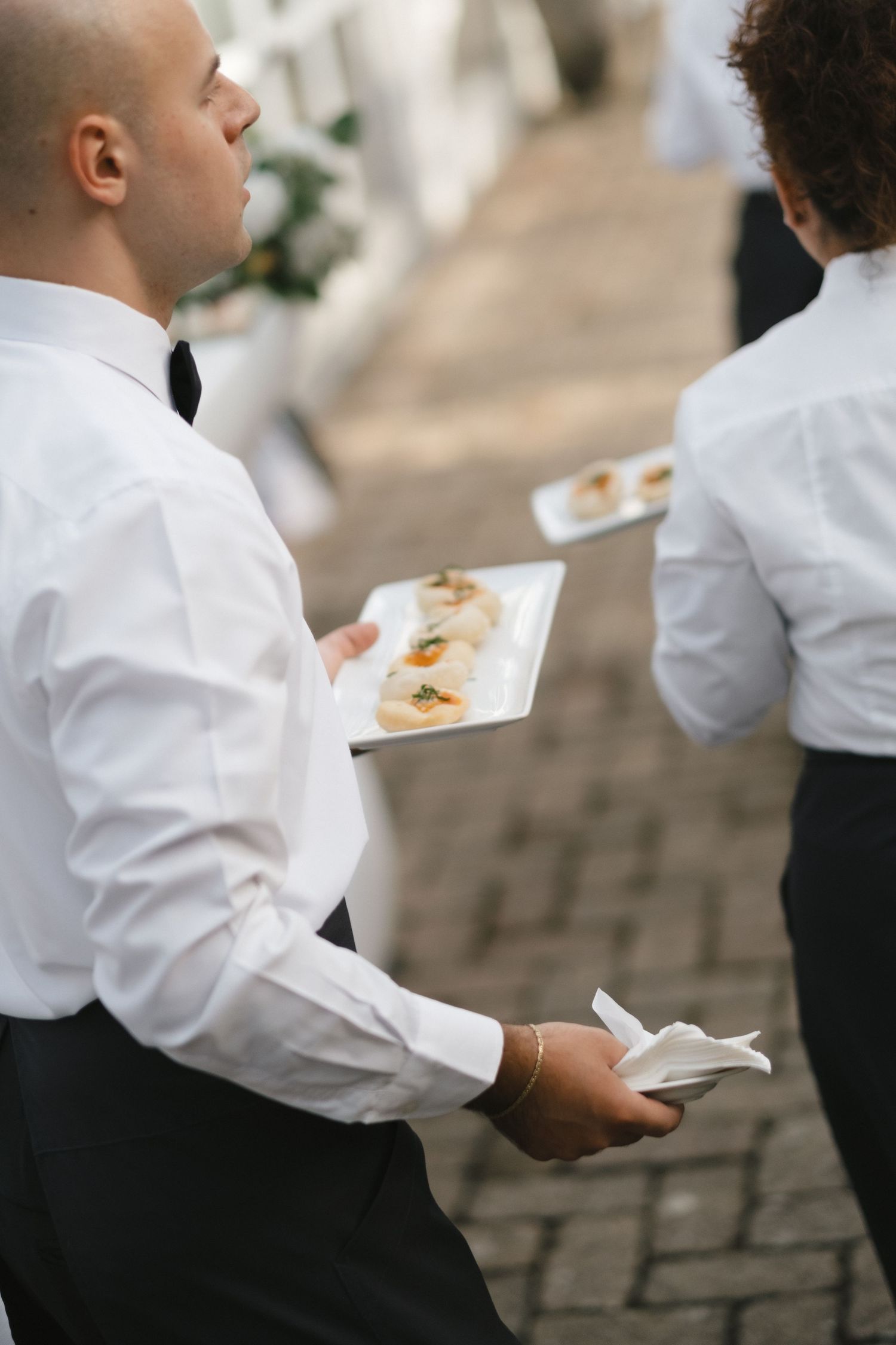 Wedding waiters in white shirts and bow ties serving canapes on white plates at an outdoor reception.