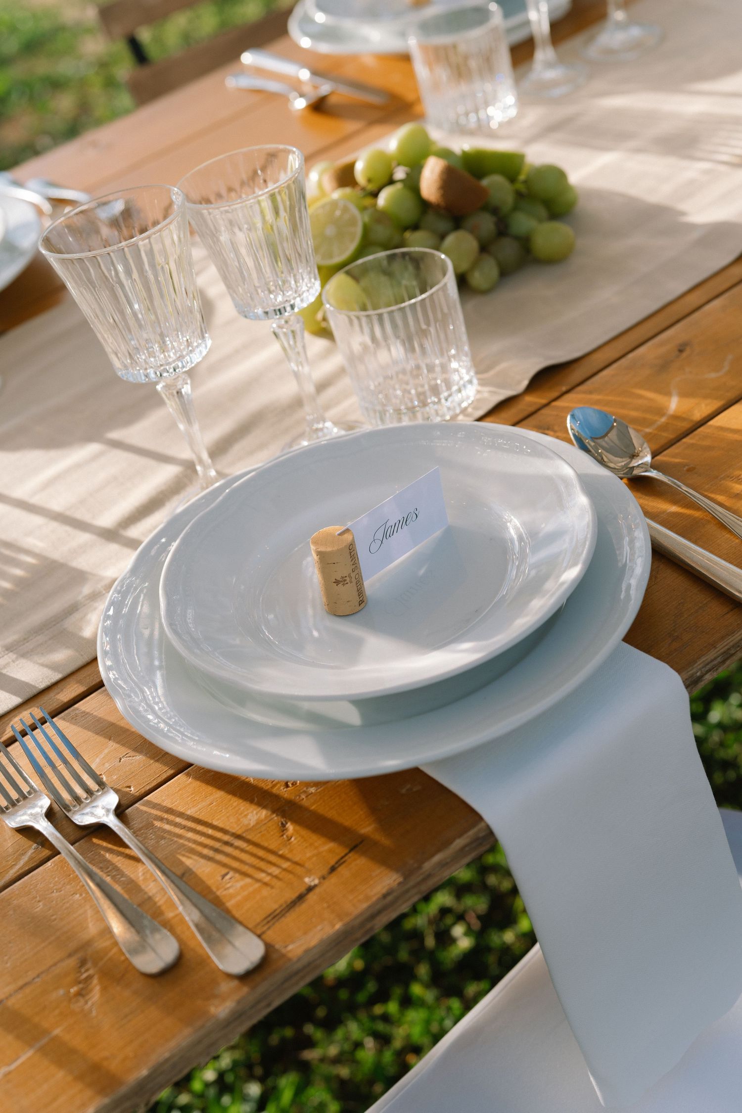 Elegant outdoor wedding table setting with white plates, crystal glasses, green grapes, and a cork place card holder.