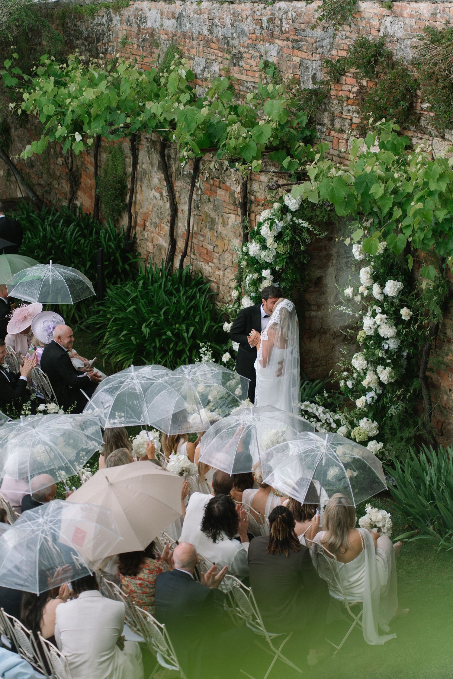 Outdoor wedding ceremony in lush garden with guests holding clear umbrellas as couple exchanges vows at floral arch.
