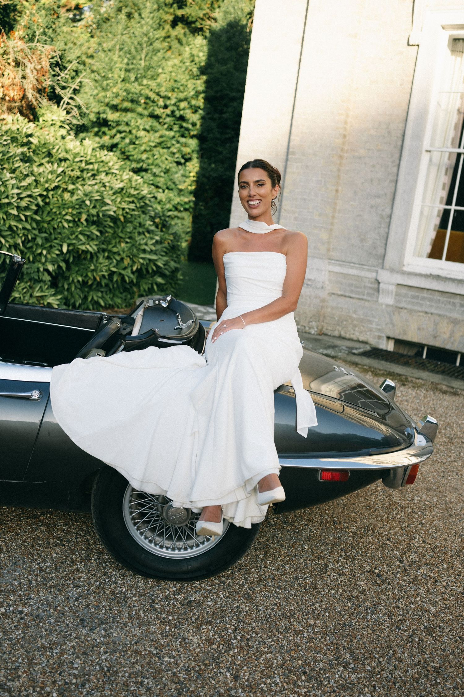 Bride in white strapless gown sitting on vintage black convertible car outside elegant estate.