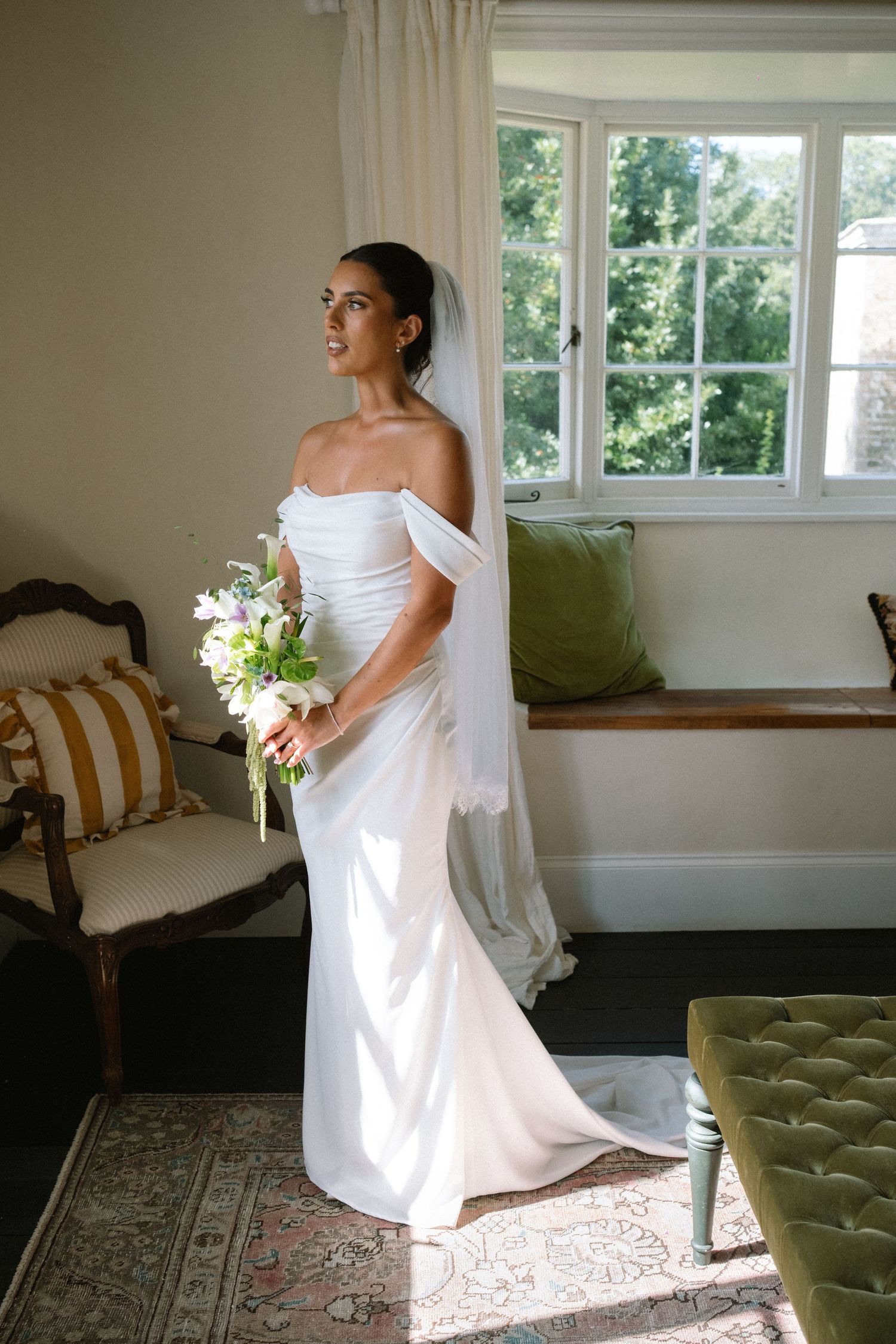 Bride in off-shoulder white gown holds floral bouquet by sunlit window in elegant room.