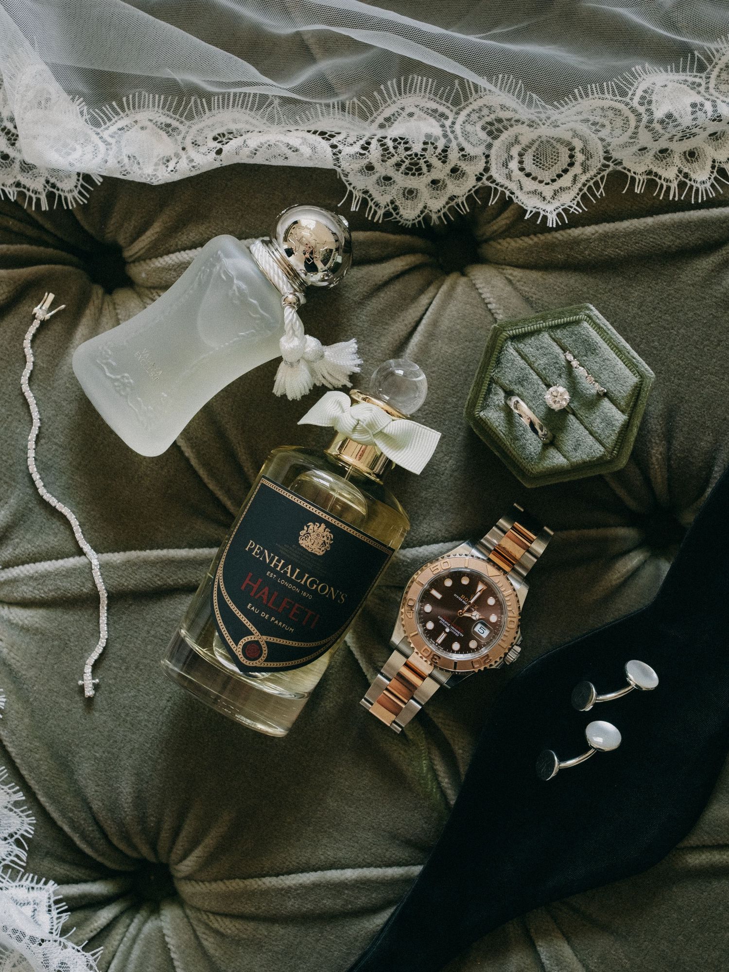 Penhaligon's perfume bottle, Rolex watch, ring box, and cufflinks arranged on green velvet with lace.
