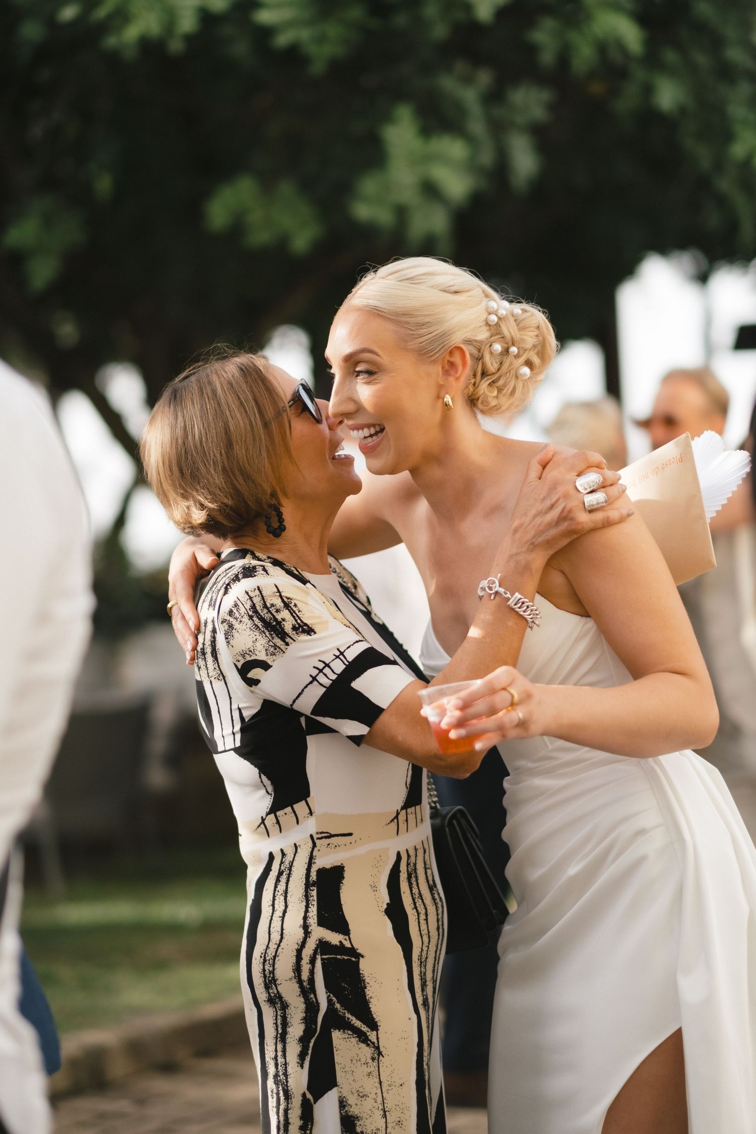 Bride in white dress embraces older woman in black and white print dress at wedding reception outdoors.