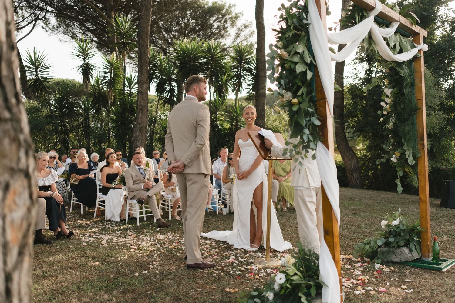 Couple exchanges vows at outdoor wedding ceremony with wooden arch and seated guests among lush green trees.