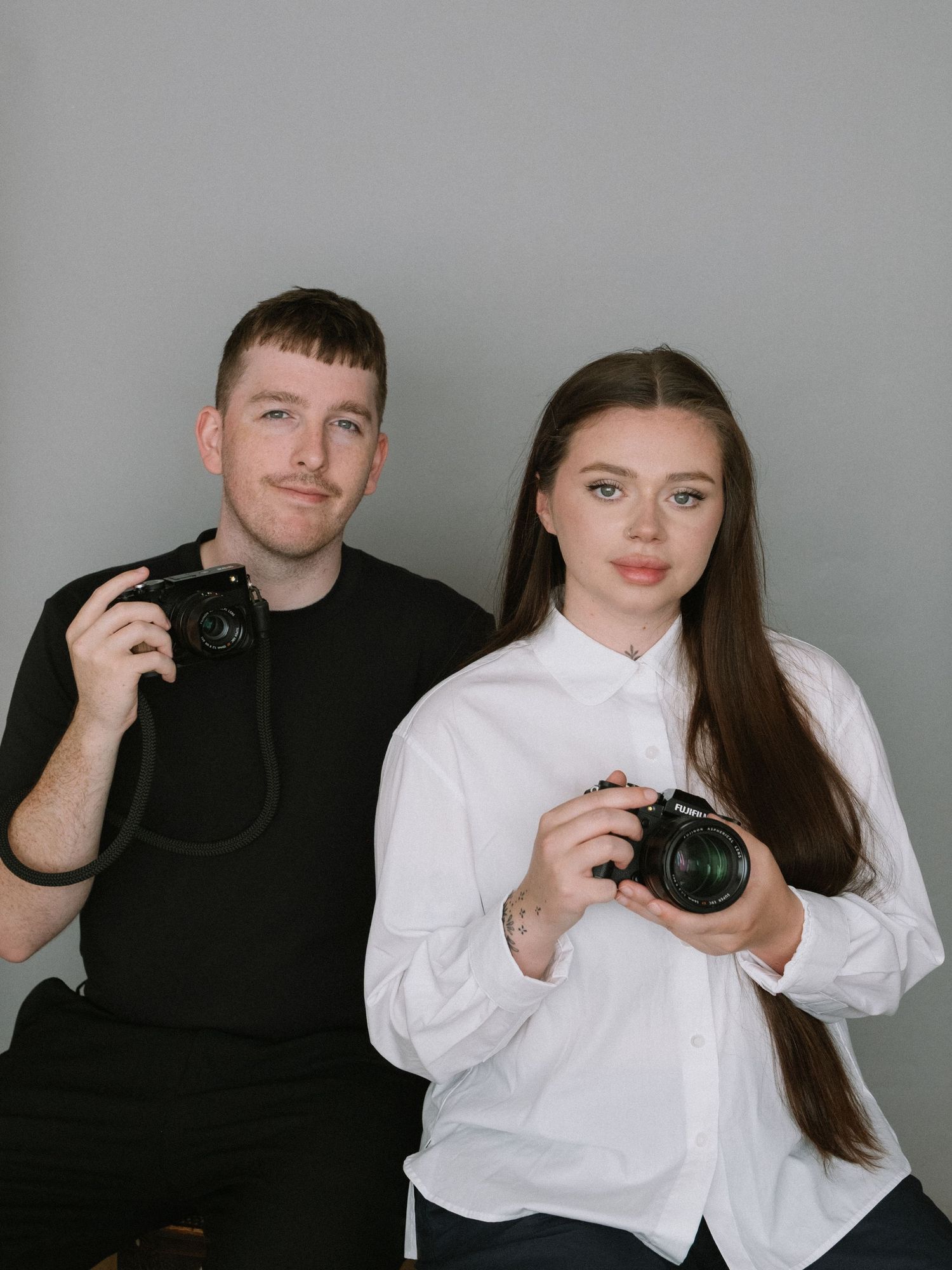 Male and female photographers holding DSLR cameras pose together in a grey studio setting.