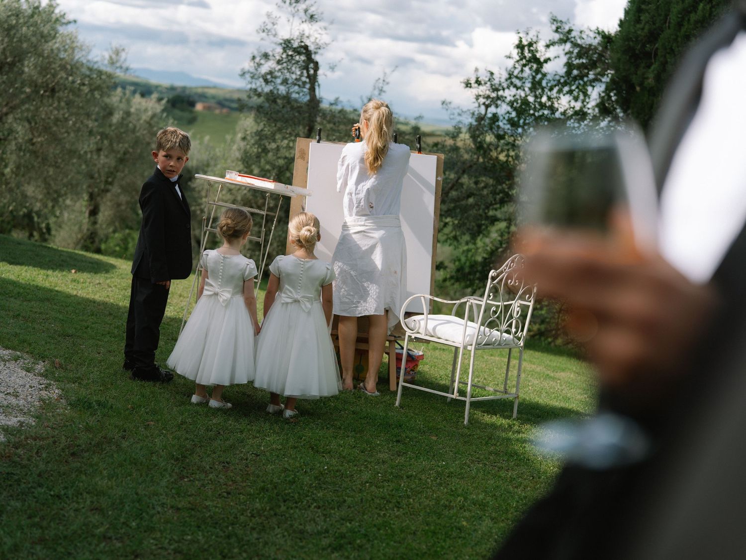 Bride in white dress stands with children in formal wear at an outdoor Tuscan wedding ceremony.