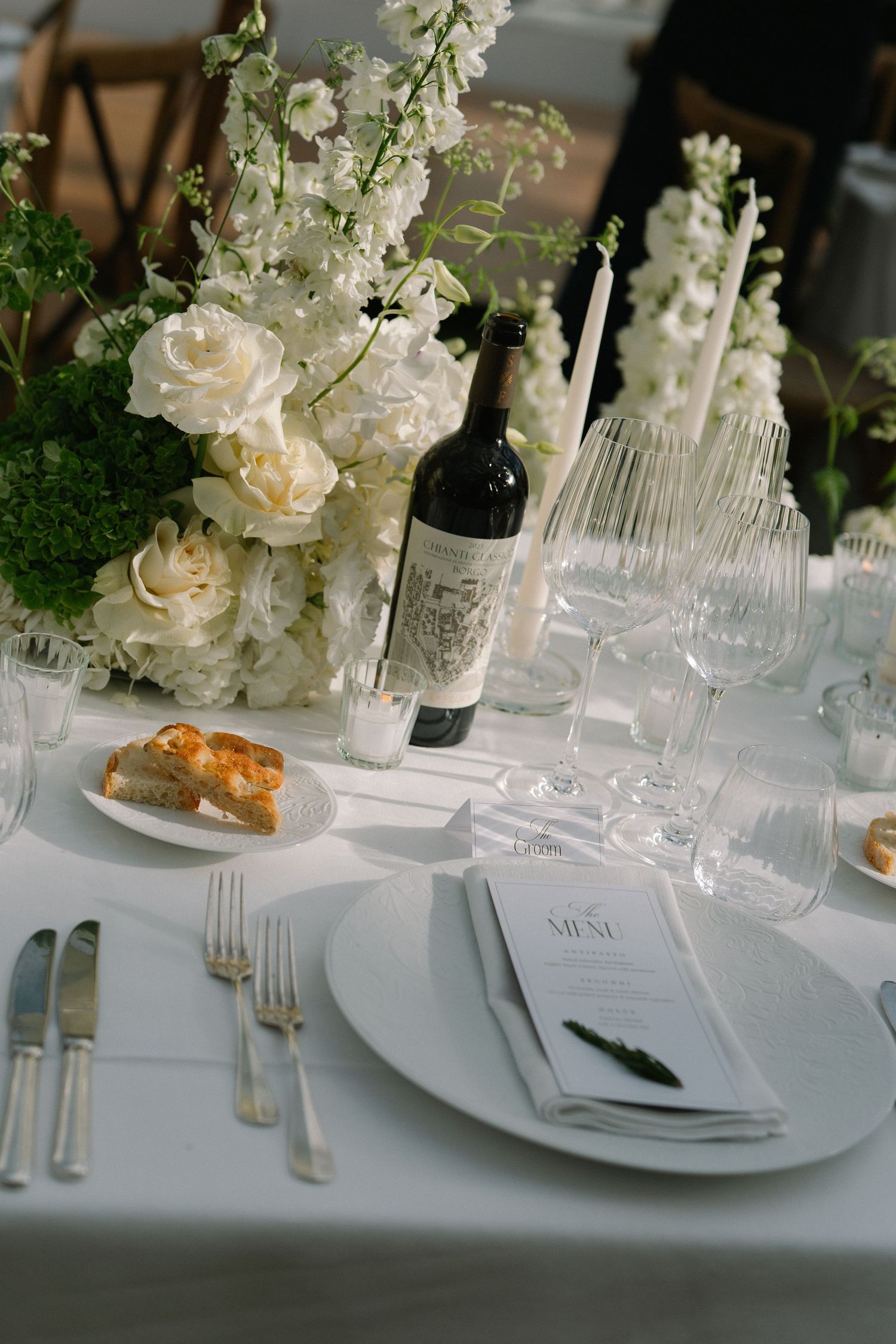 Elegant wedding table setting with white roses, delphinium, crystal glasses, and Italian wine bottle.