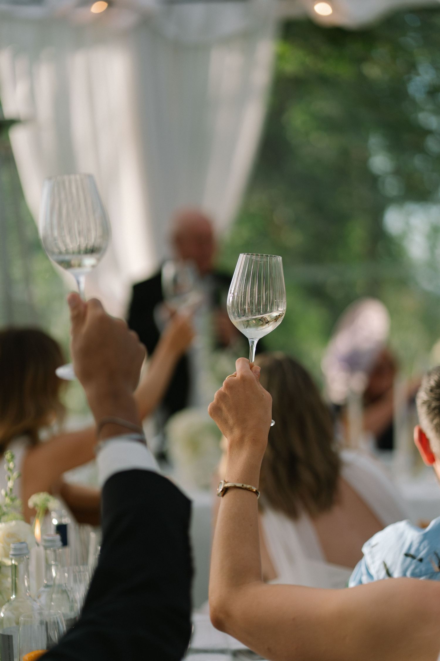 Wedding guests raising crystal wine glasses in a toast during a reception speech inside a clear tent venue.