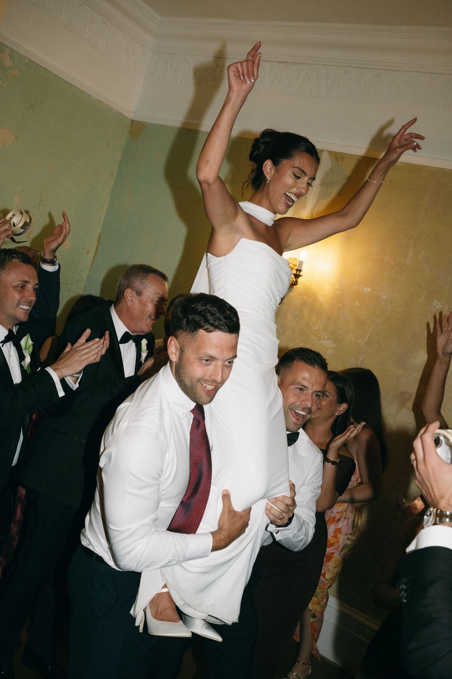 Bride in white gown lifted on groomsman shoulders celebrating at wedding reception with guests dancing.
