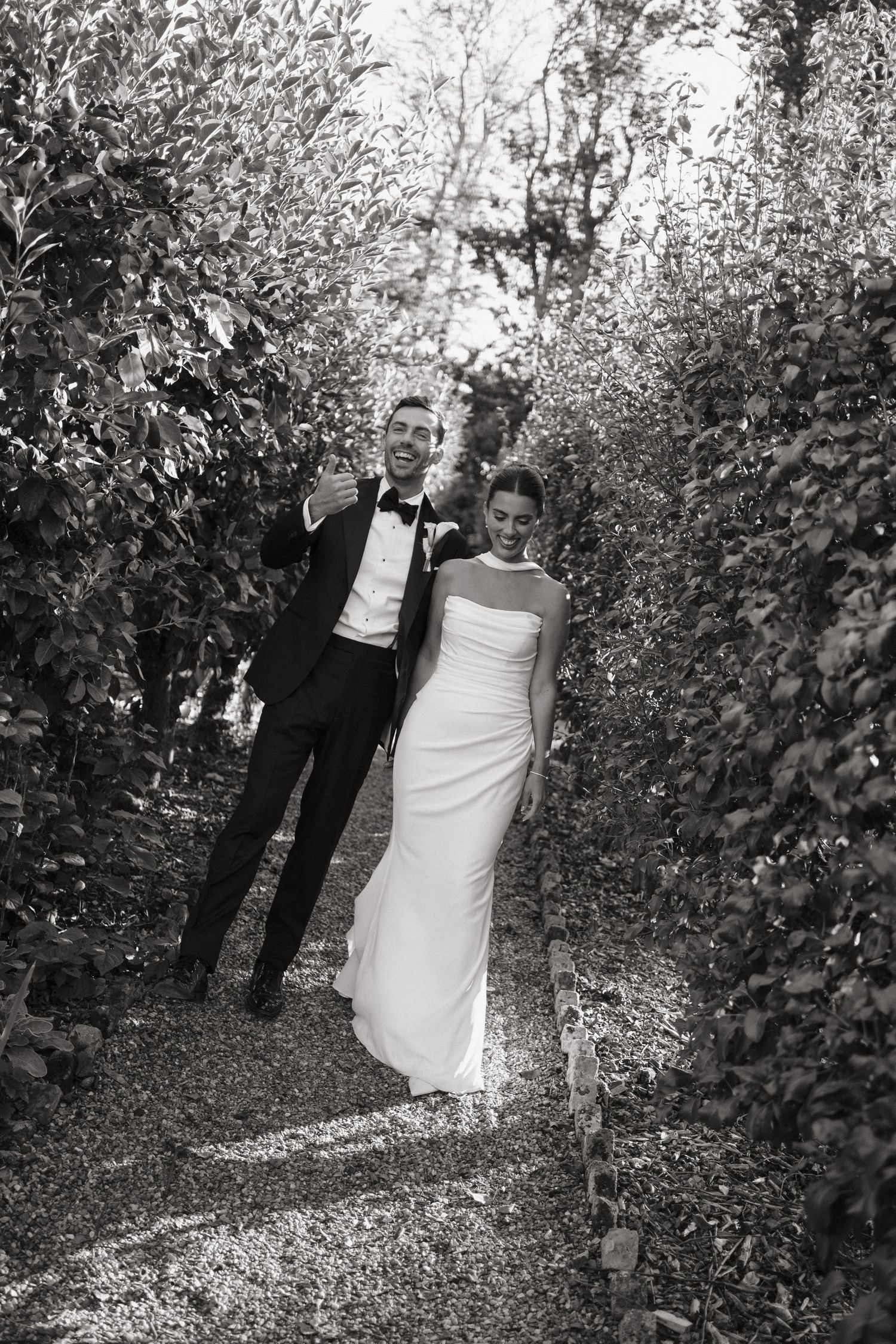 Black and white photo of a bride and groom walking through a lush garden path on their wedding day.