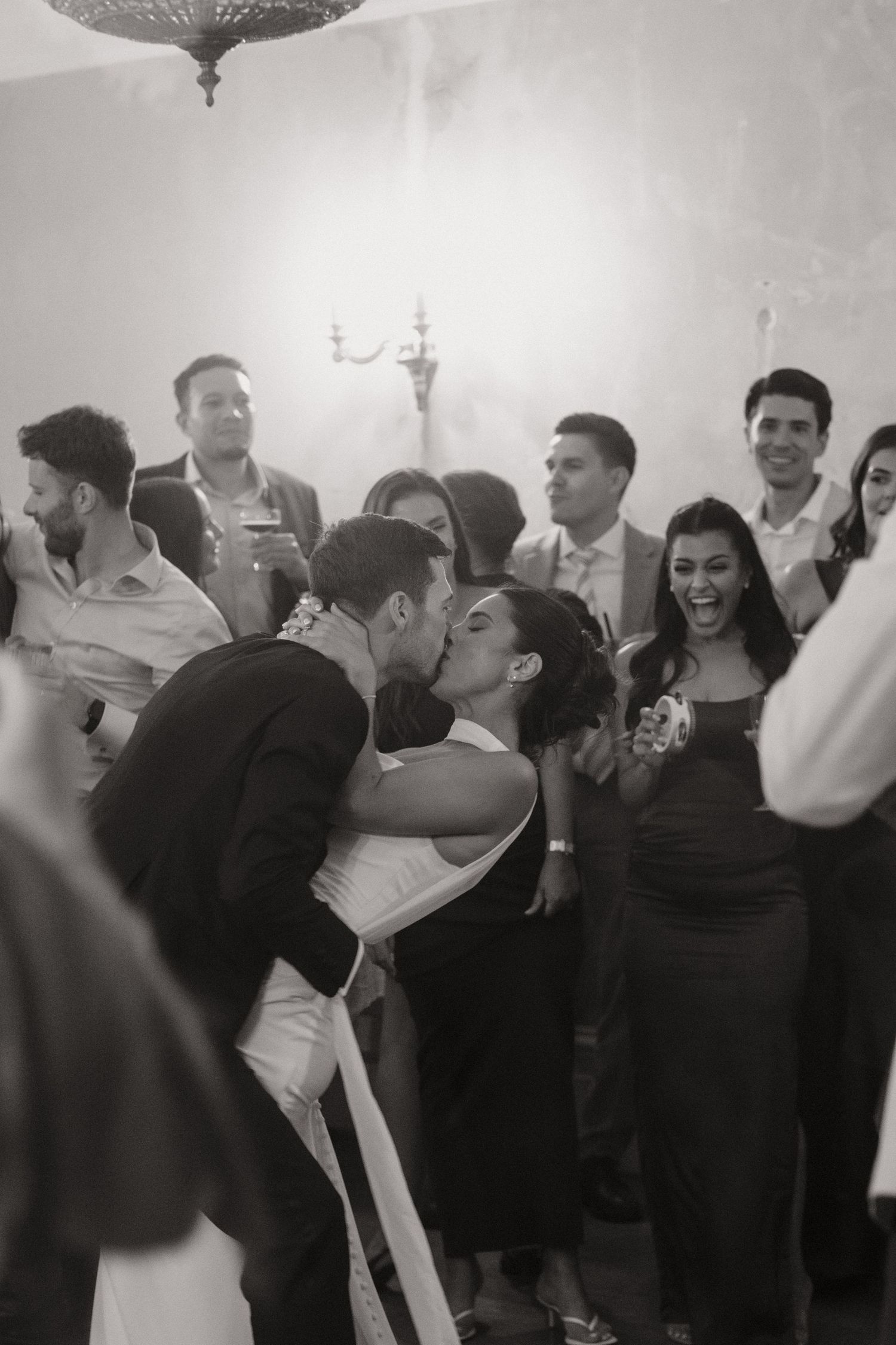Black and white photo of a couple sharing a kiss at their wedding reception surrounded by smiling guests.