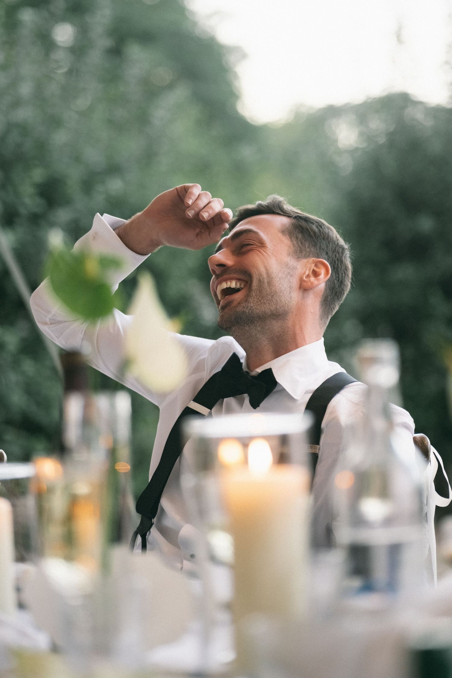 A laughing groom in a white shirt, bow tie, and suspenders enjoys a joyful moment at an outdoor wedding reception.