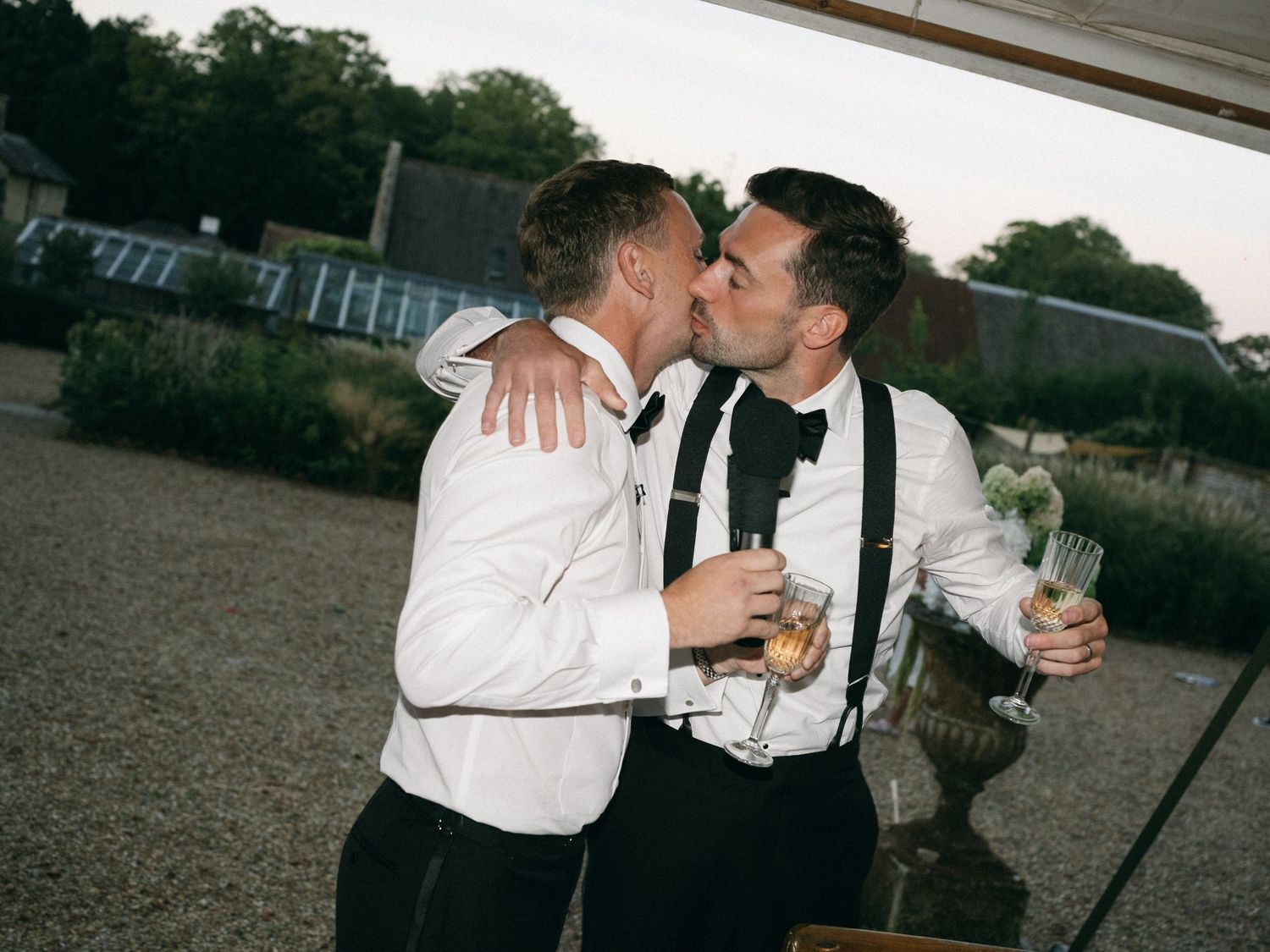 Two grooms in formal attire sharing a kiss at their outdoor wedding reception while holding champagne glasses.