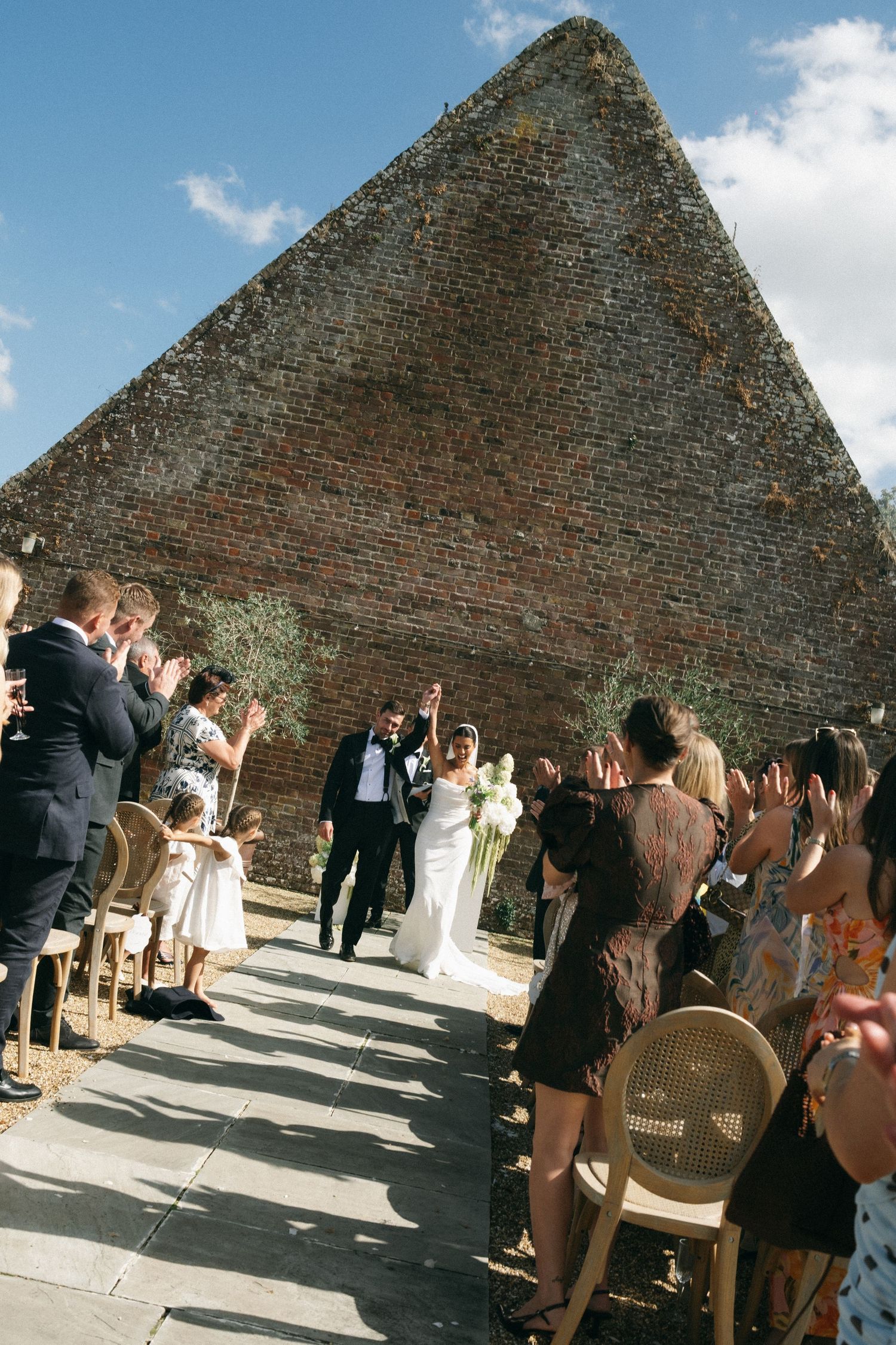 Bride and groom walk down outdoor aisle at brick pyramid venue, guests applauding on sunny wedding day.