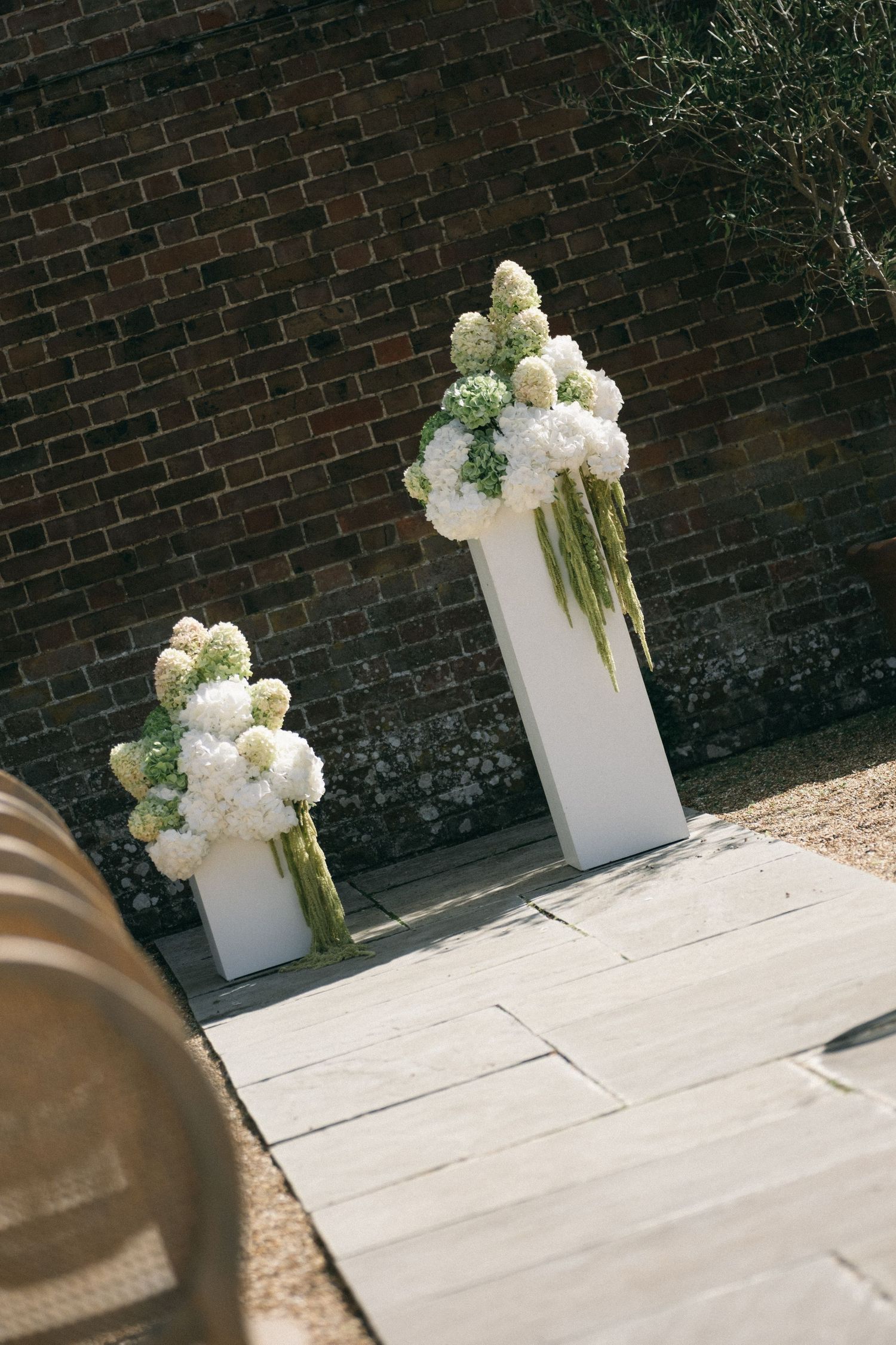 Two white pedestal stands with lush white hydrangea and green floral arrangements against a rustic brick wall outdoor setting.