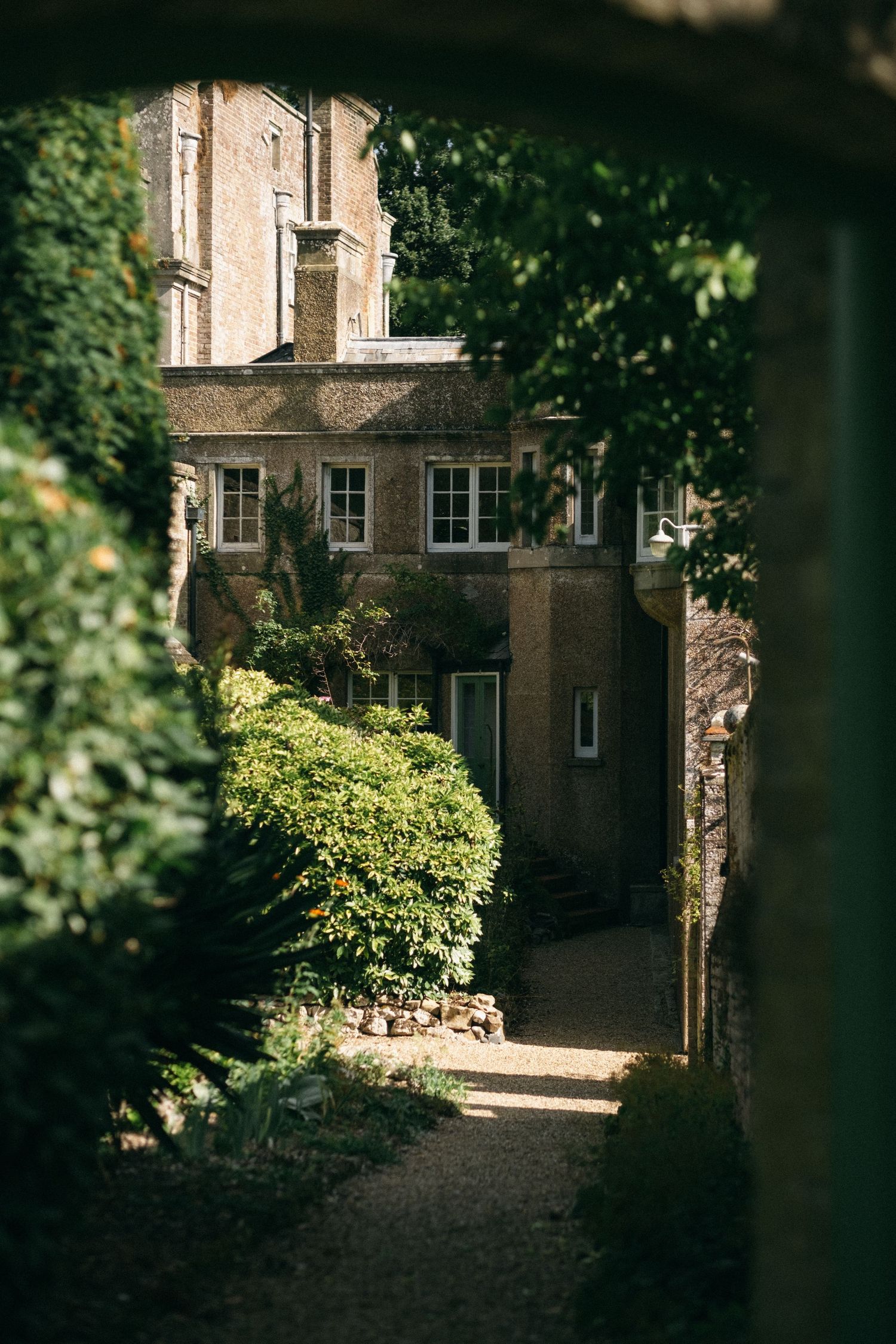 Charming English manor house viewed through lush garden archway, with gravel path and manicured hedges in warm sunlight.