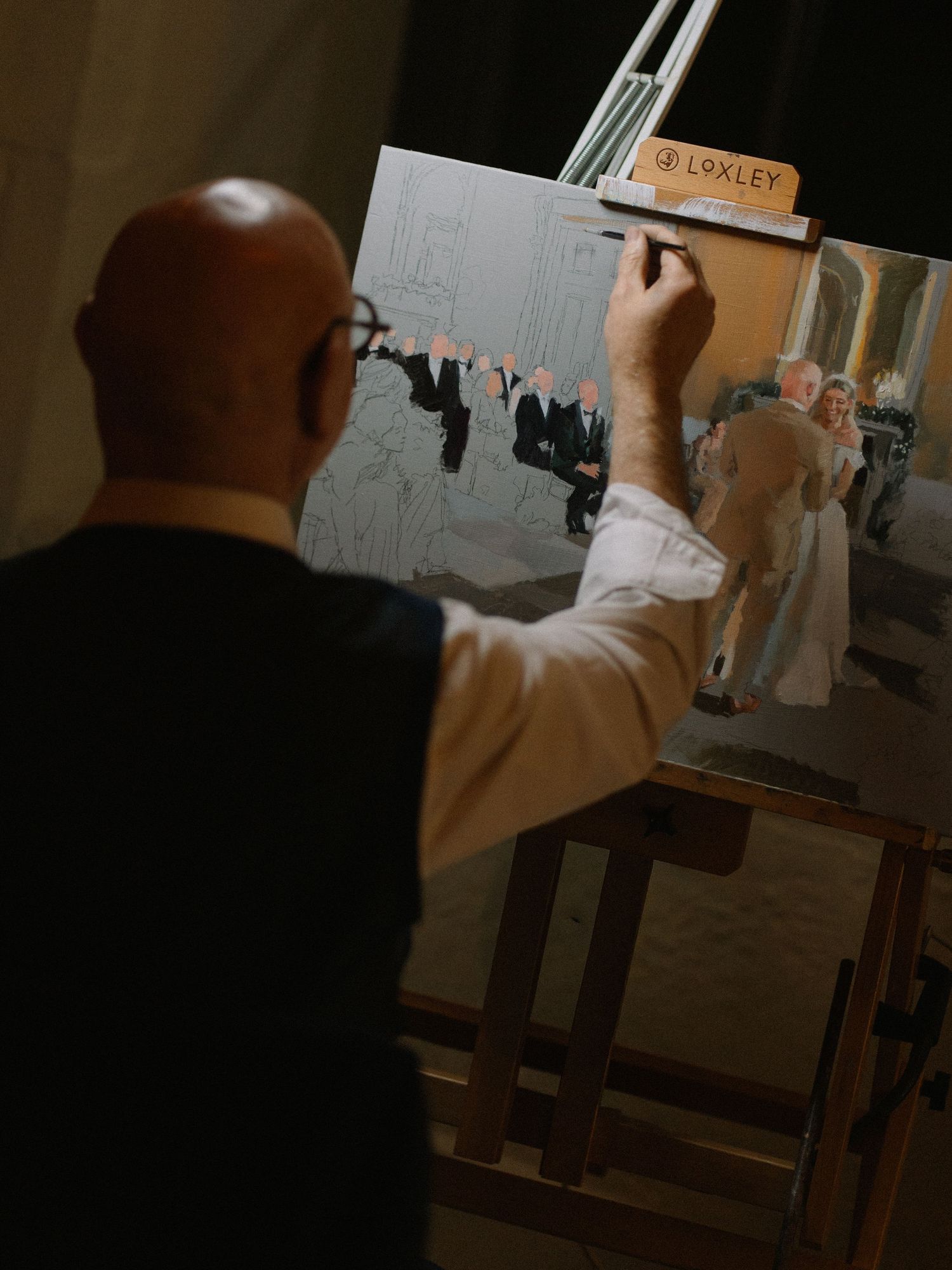 Bald artist painting a live wedding scene on a Loxley easel, capturing guests in formal attire at an elegant event.