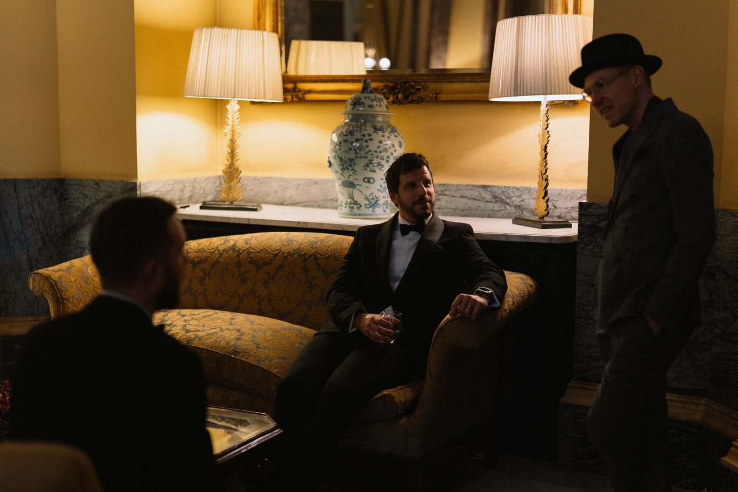 Three men in formal attire converse in an elegantly lit room with classic decor and warm ambient lighting.