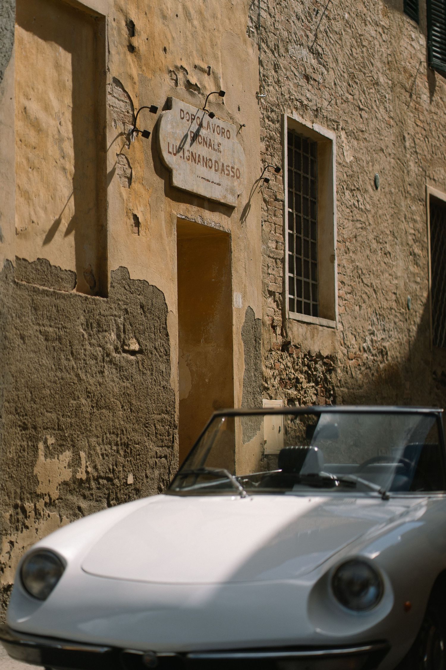 Vintage white Alfa Romeo Spider parked beside a weathered stone building in a charming Italian village.
