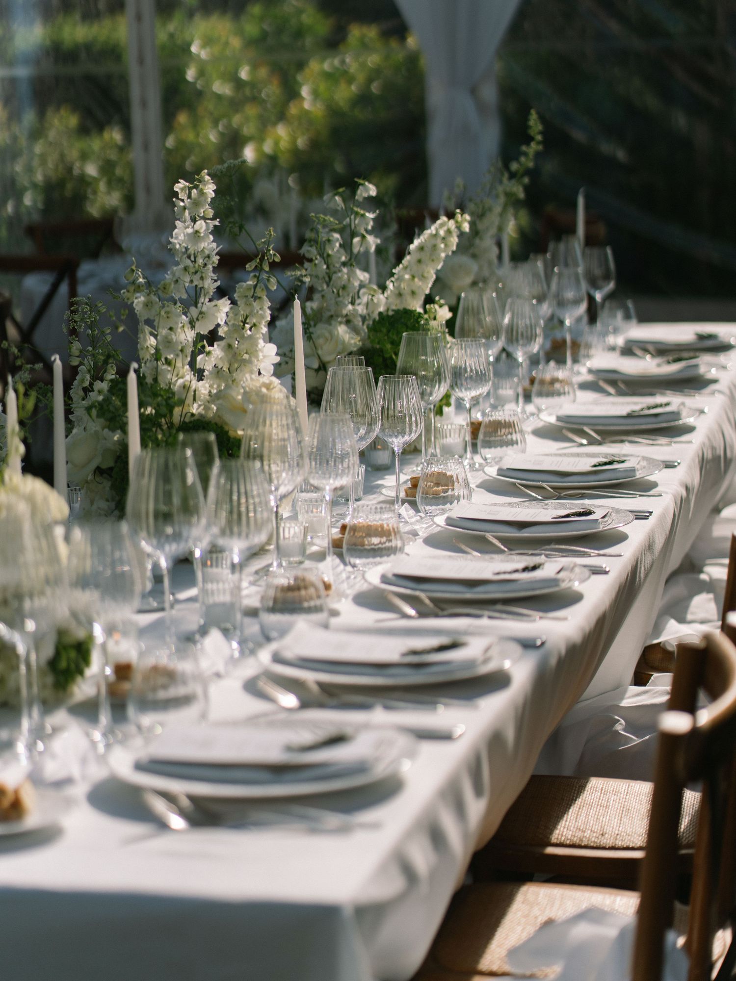 Elegant wedding reception long table with white floral centerpieces, crystal glassware, and white linen under a clear tent.