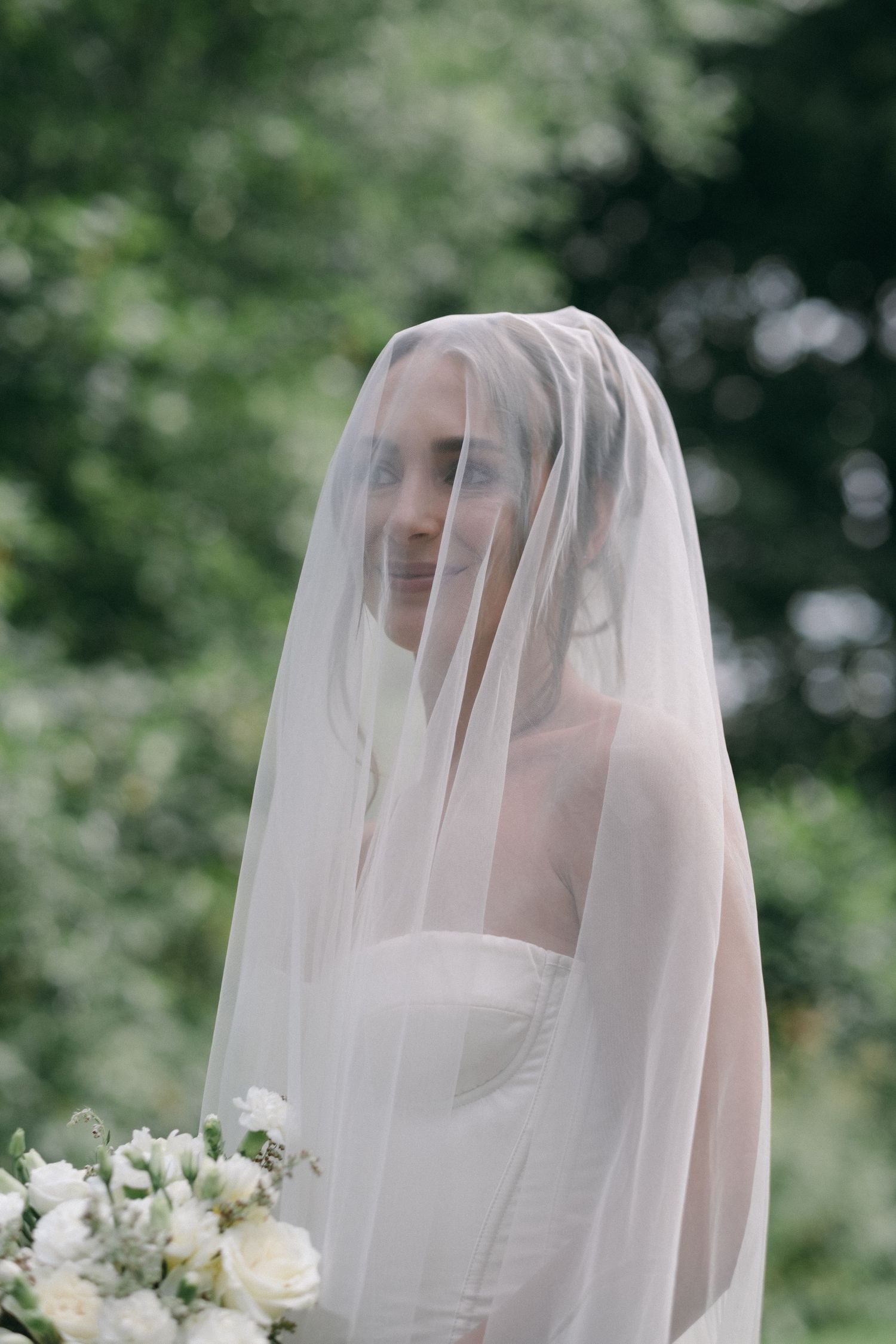 Bride smiles softly beneath a sheer wedding veil with a white floral bouquet in a garden.