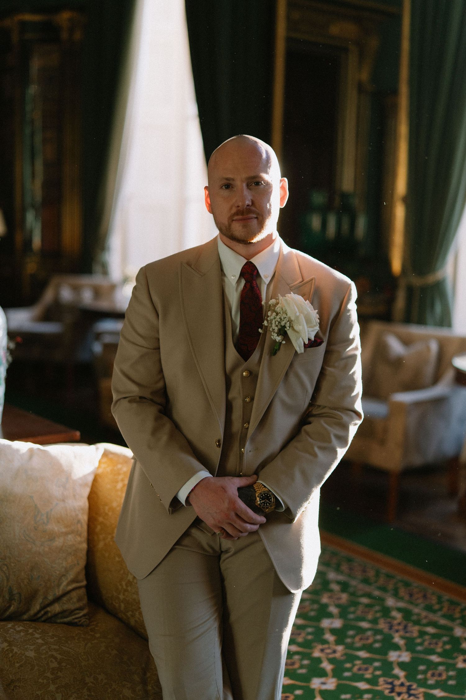 Bald groom in beige three-piece suit with white boutonniere and burgundy tie standing in elegant green room.
