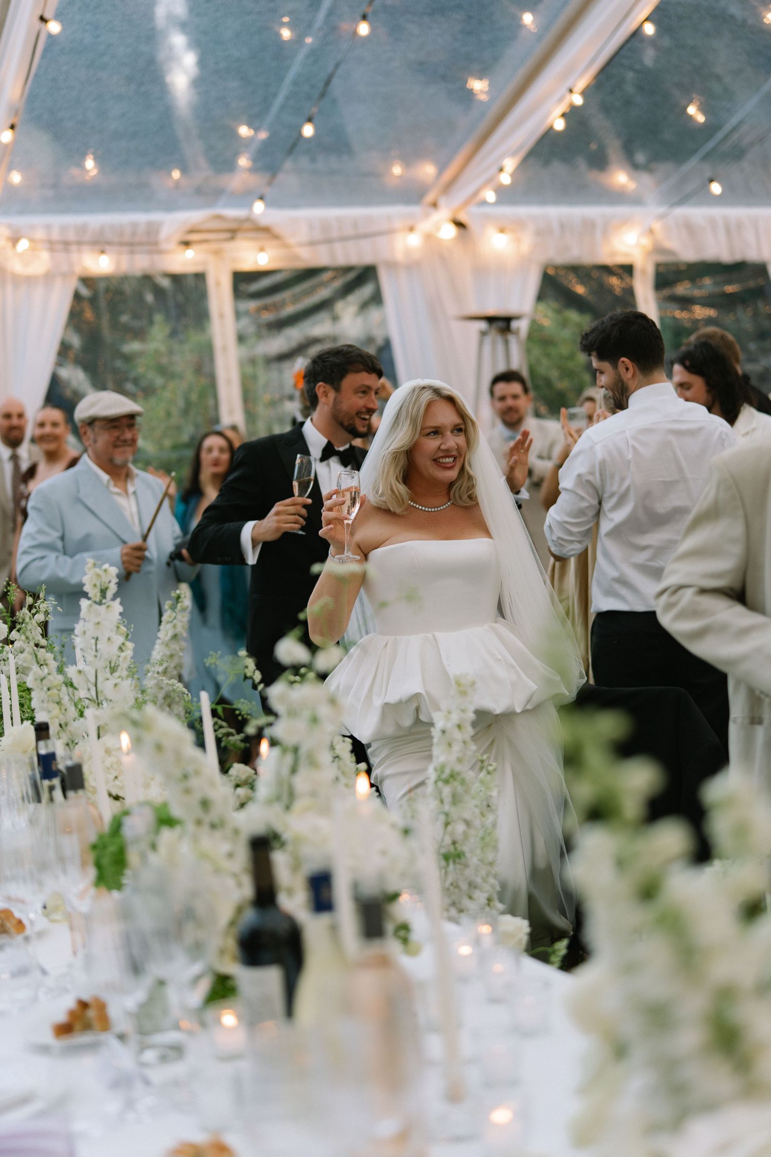 Bride in white peplum gown raises champagne glass at elegant tented wedding reception with string lights and floral decor.