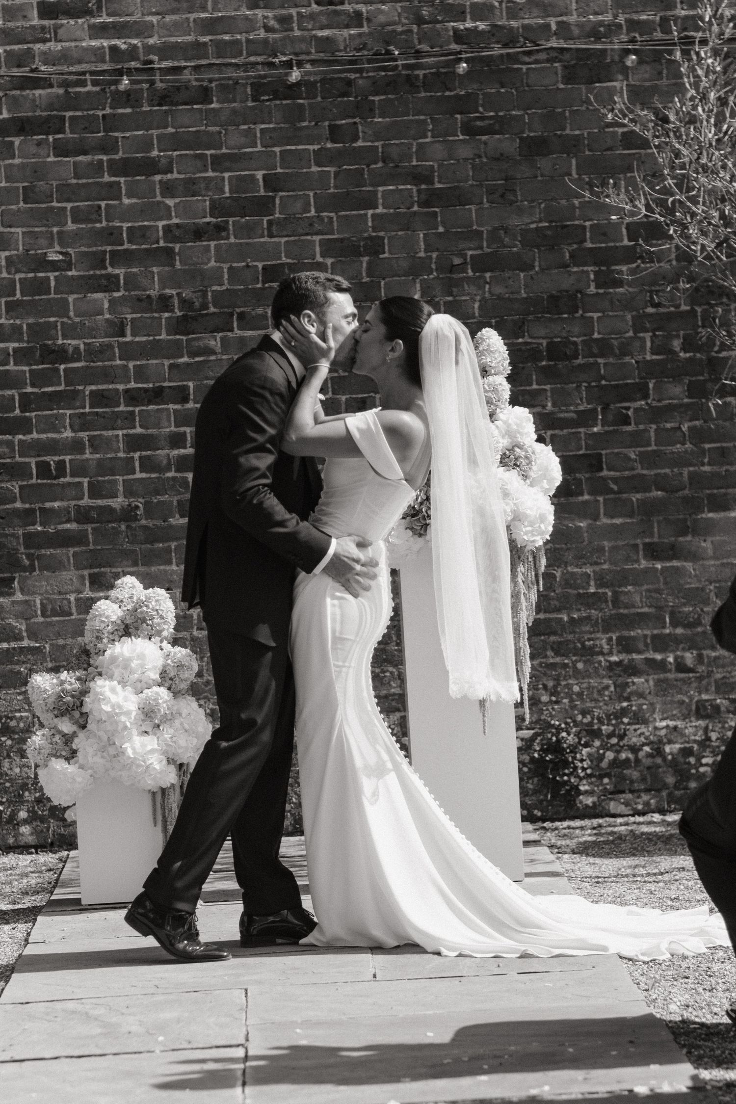 Black and white photo of a bride and groom sharing their first kiss at an outdoor wedding ceremony with floral arrangements.