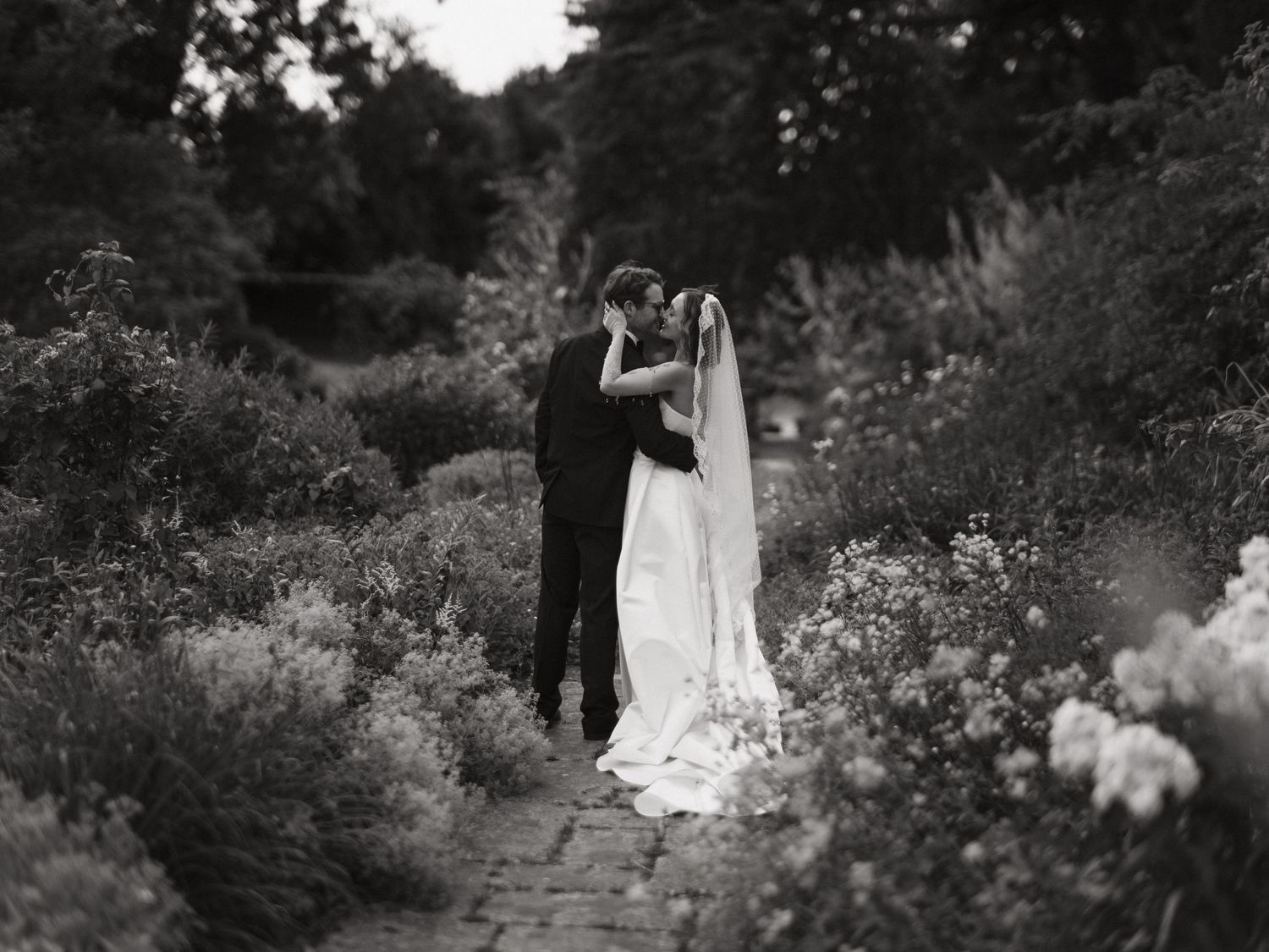 Black and white photo of a bride and groom kissing on a garden path surrounded by lush flowers and greenery.