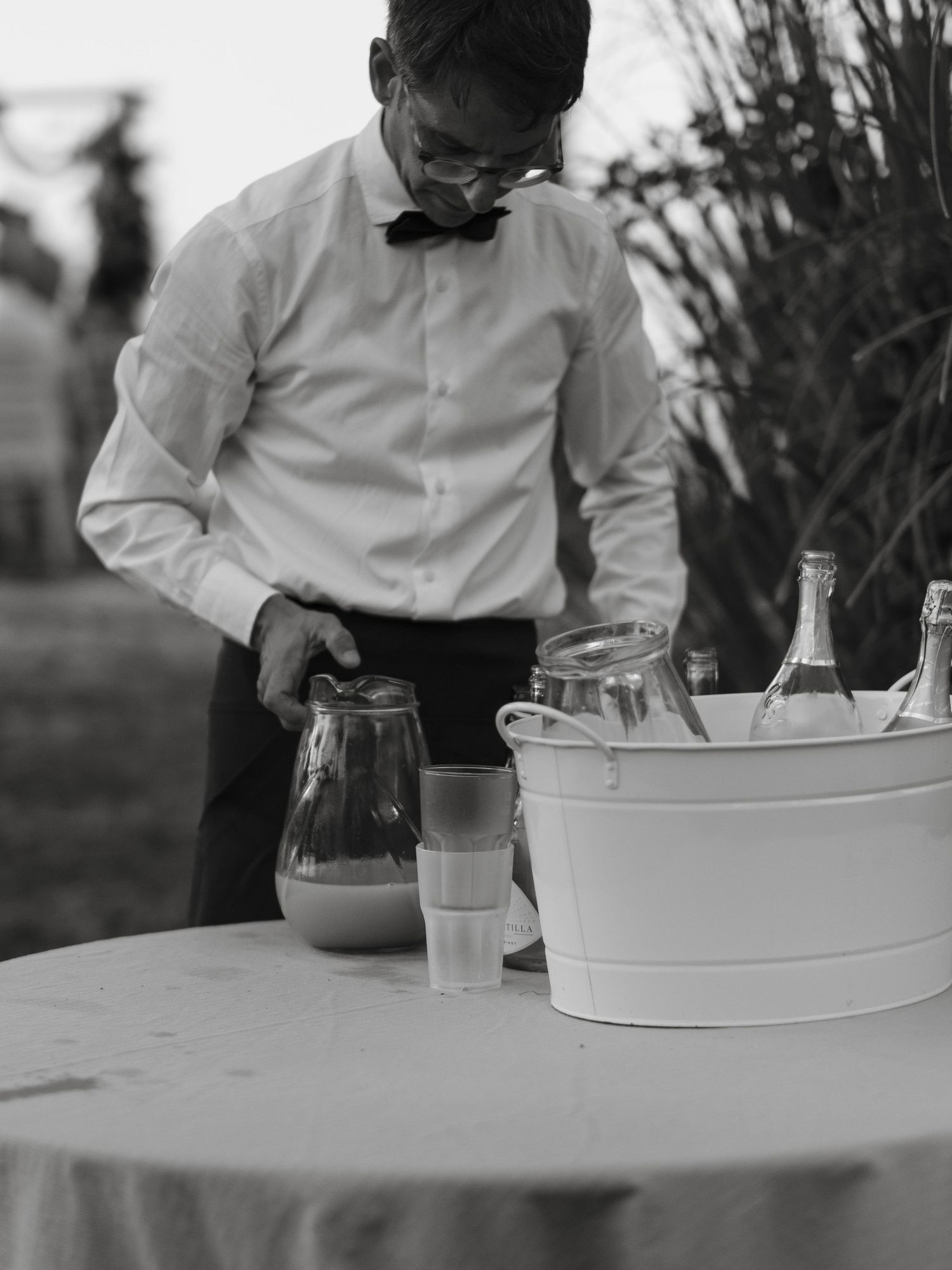 A waiter in a bow tie pours a drink at an outdoor event table with bottles in an ice bucket.