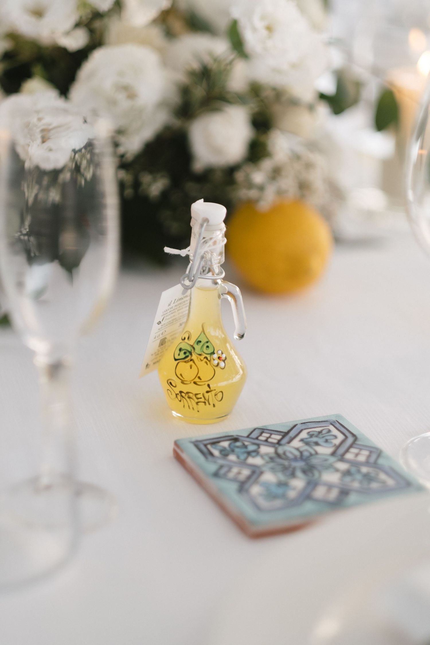 Small bottle of limoncello with hand-painted lemon design beside a decorative tile and champagne glass at a wedding reception.