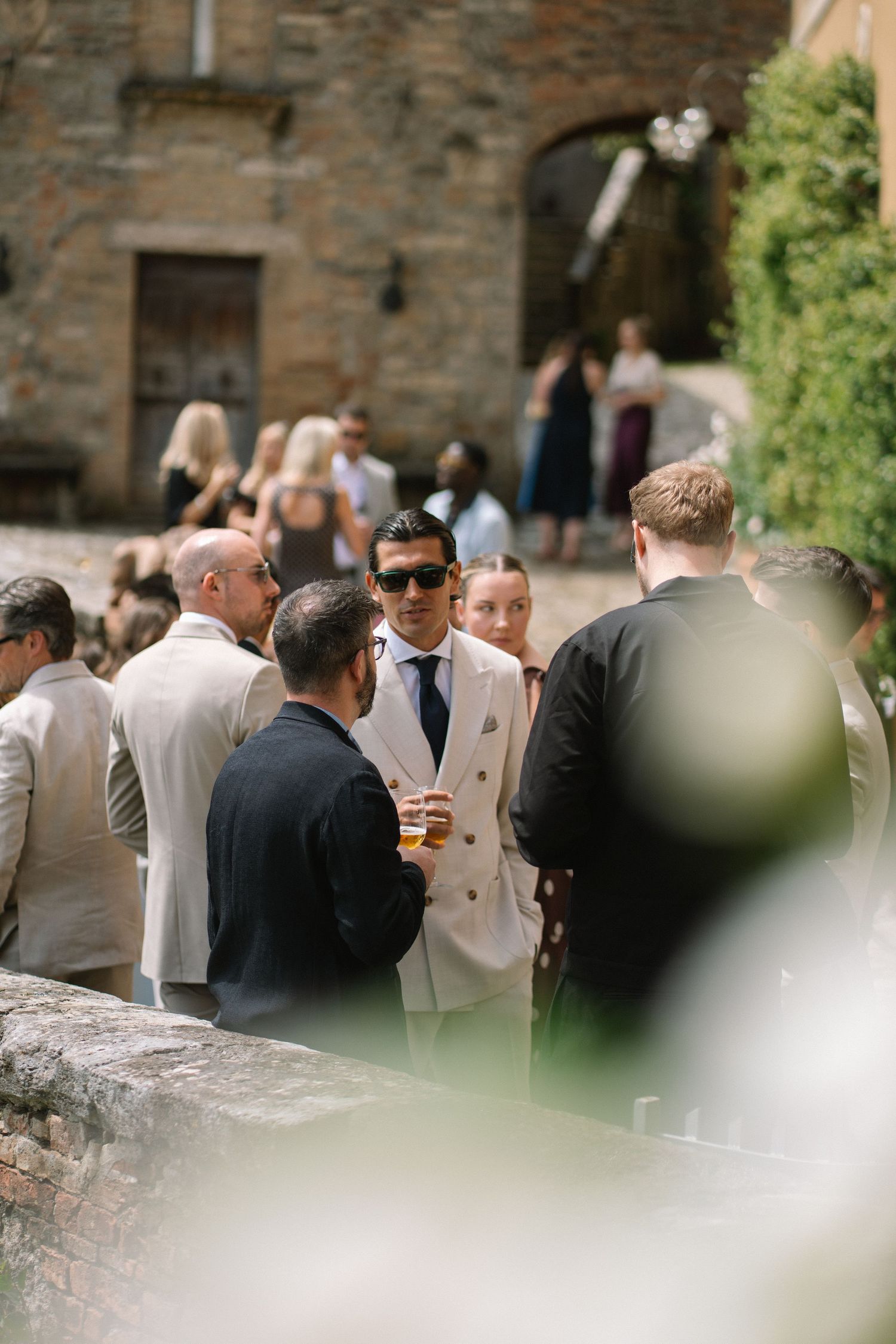 Wedding guests mingling outdoors at rustic stone Italian villa in summer sunshine.