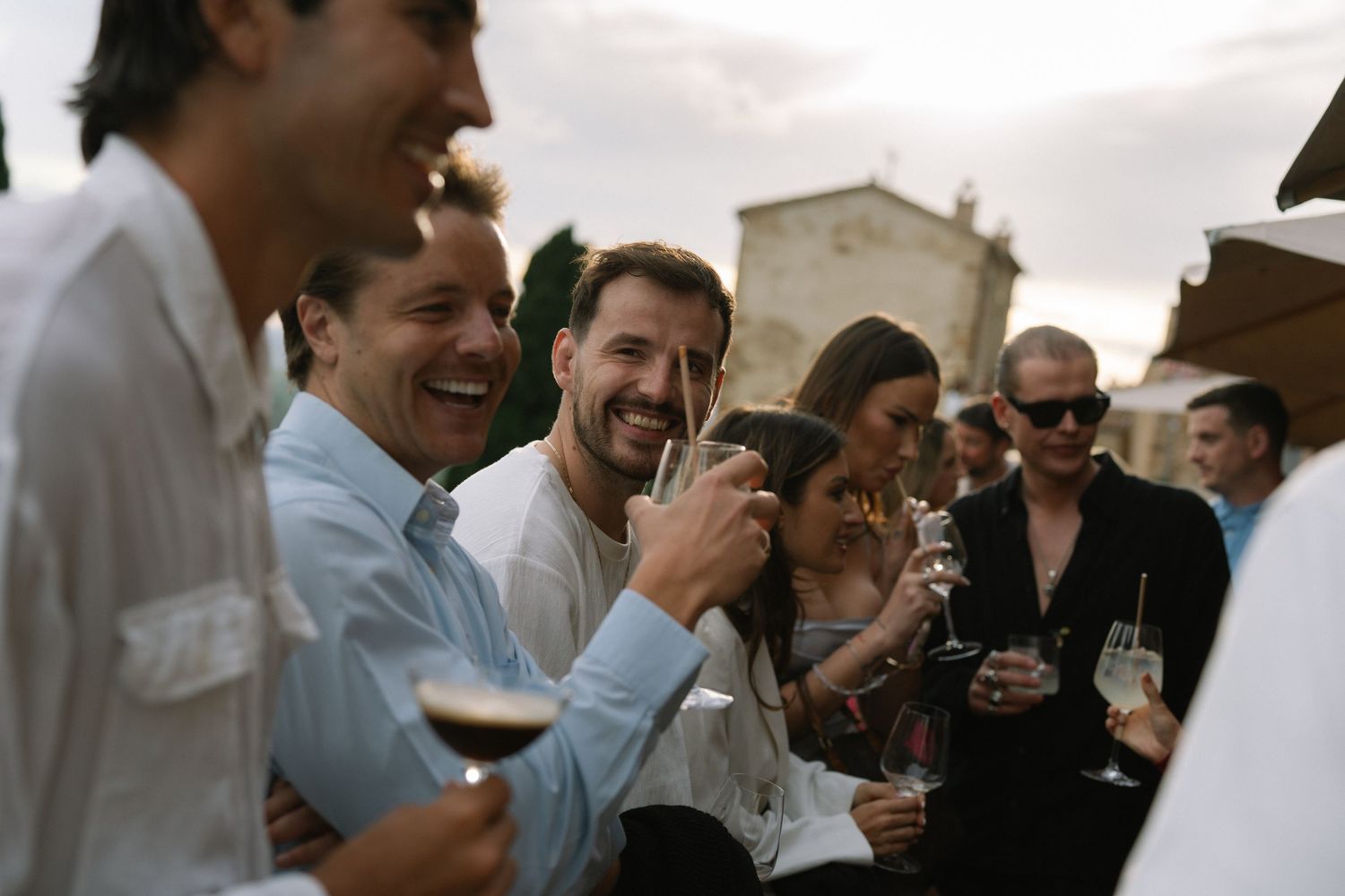 Group of cheerful wedding guests laughing and drinking champagne at an outdoor Tuscan wedding celebration.