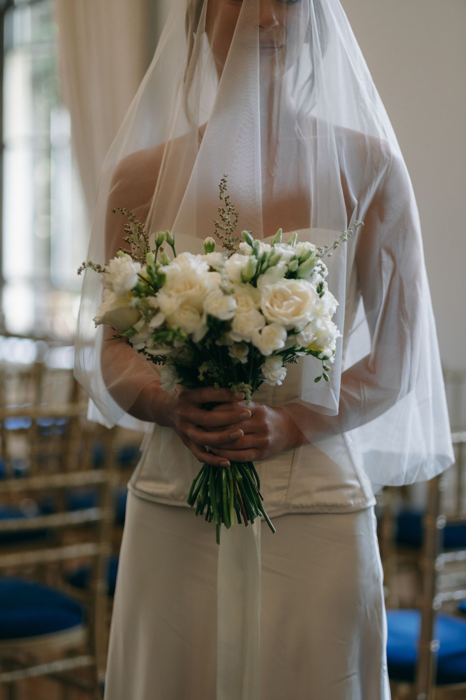 Bride in white gown and veil holding elegant bouquet of white roses and greenery at wedding ceremony.