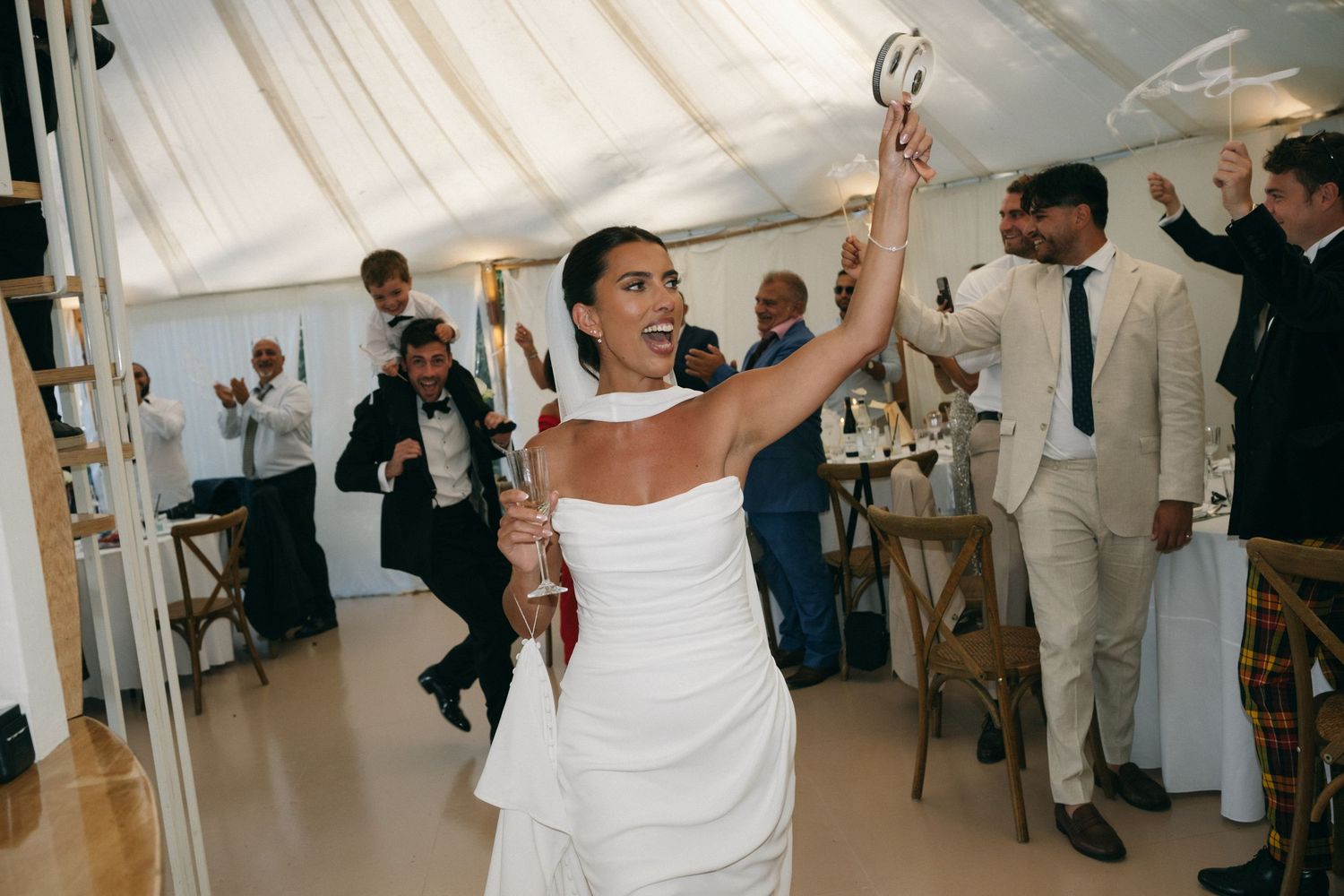 Bride in white dress dancing joyfully at wedding reception, raising hand with guests celebrating in marquee tent.