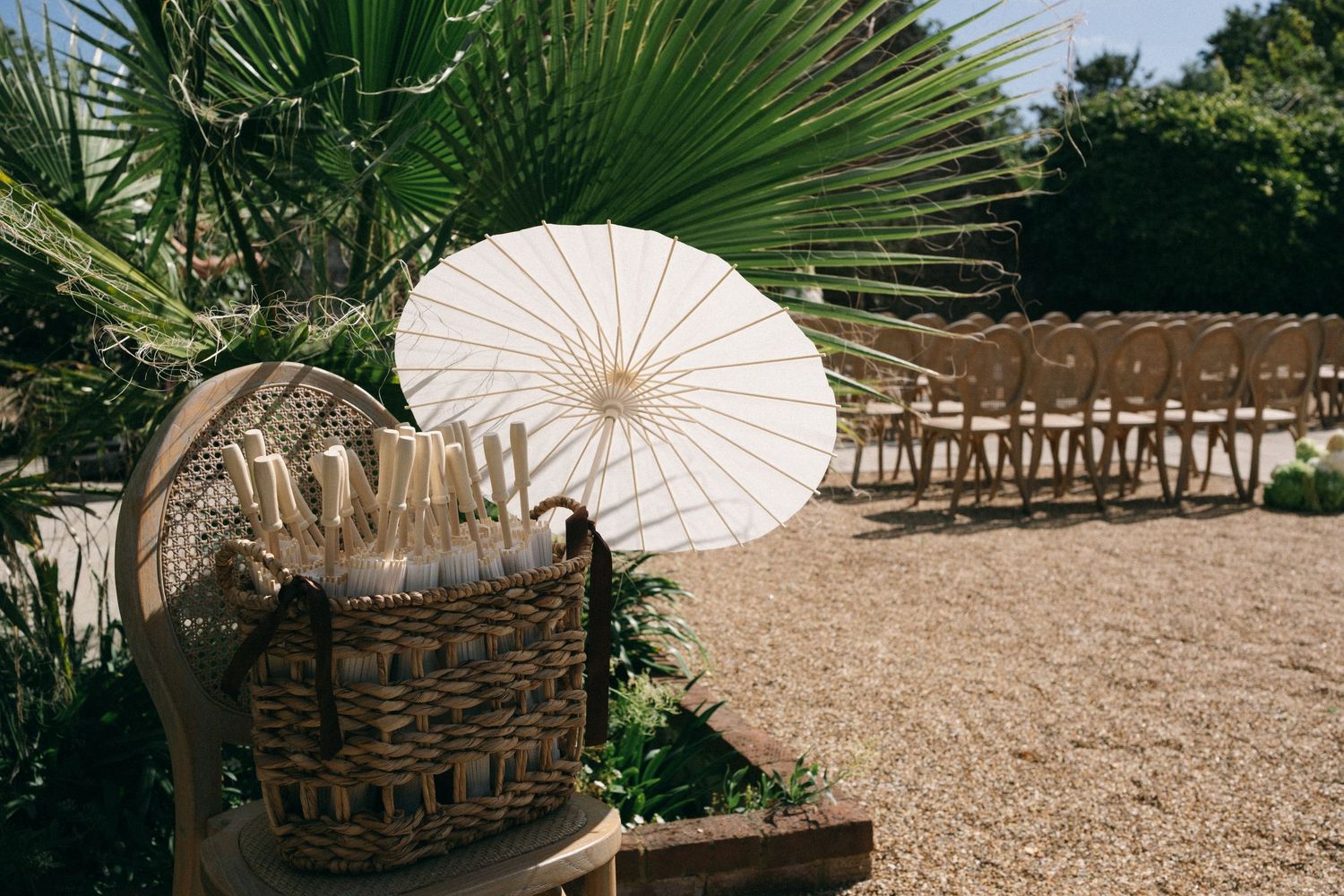 Outdoor wedding ceremony setup with wicker basket, white parasol, and wooden chairs on gravel surrounded by tropical palms.