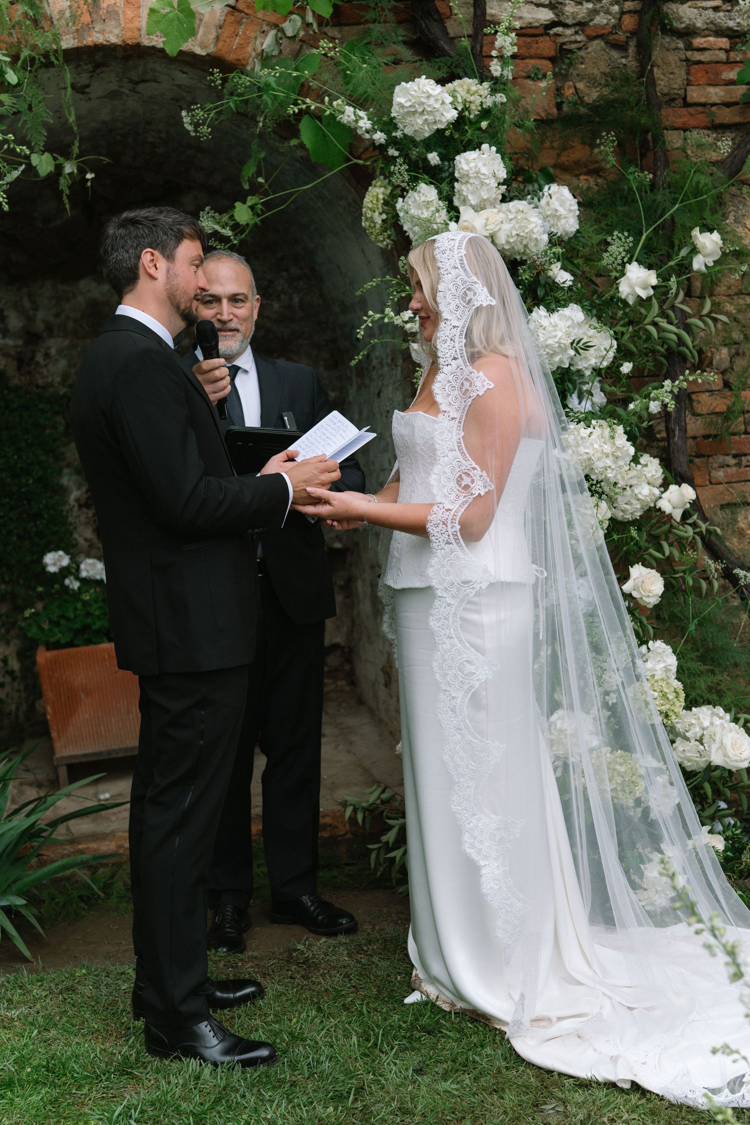 Groom reads wedding vows to bride under a white floral arch at an outdoor garden ceremony.