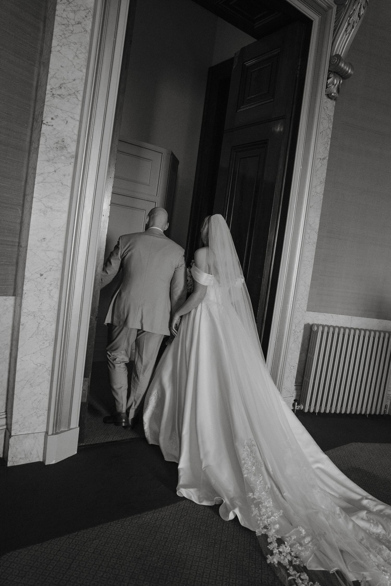 Black and white photo of bride in white gown and veil walking through elegant doorway with suited companion.