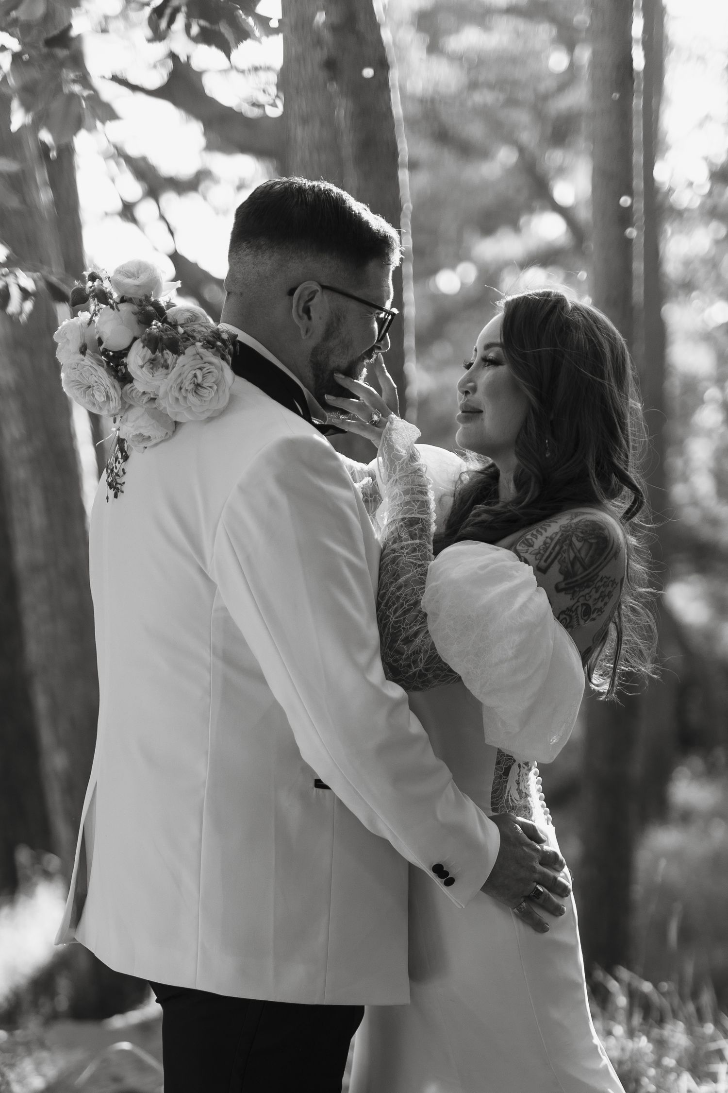 Black and white photo of a tattooed bride and groom embracing in a forest, wearing elegant wedding attire.