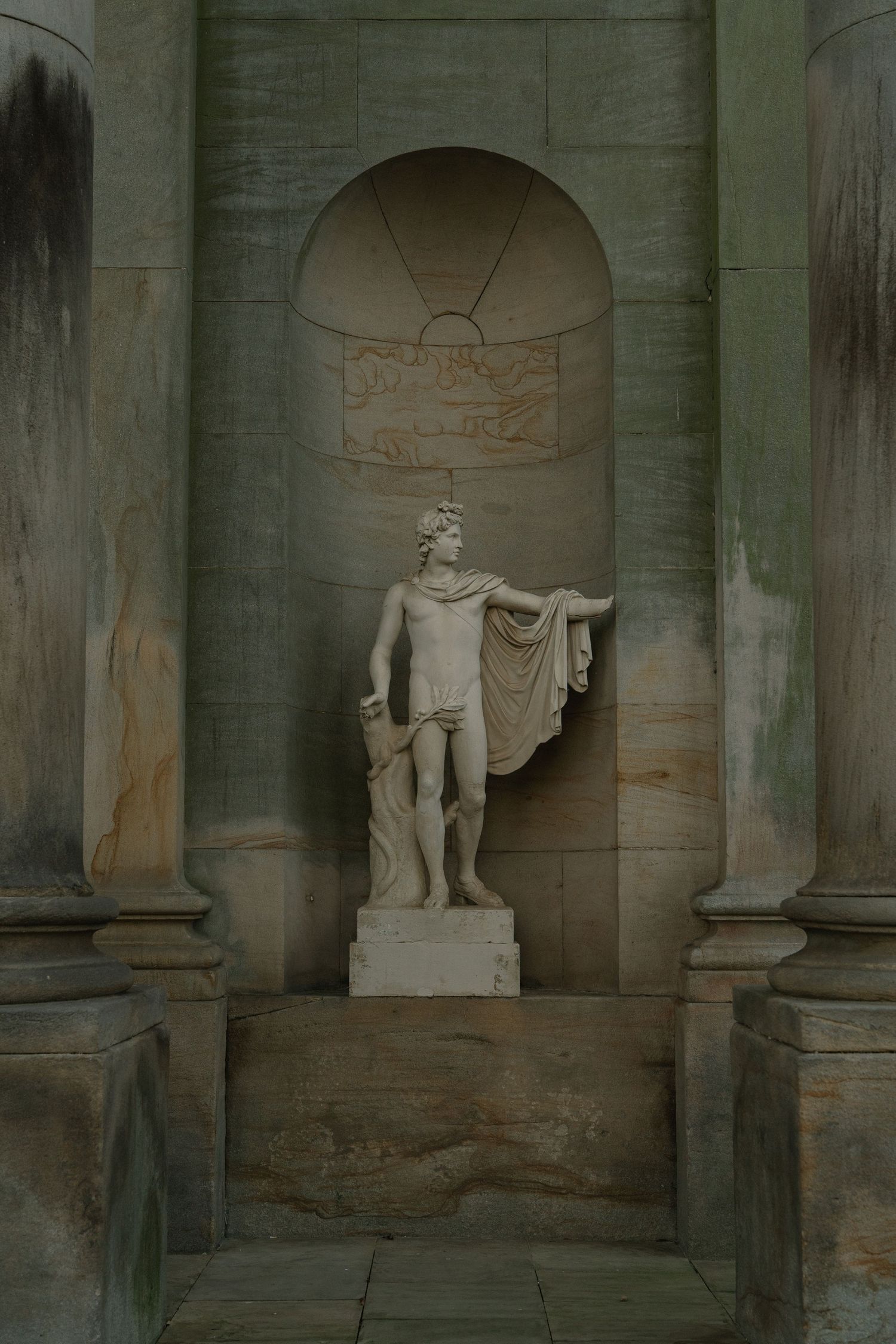 Classical marble statue of a draped male figure in a stone niche flanked by columns.