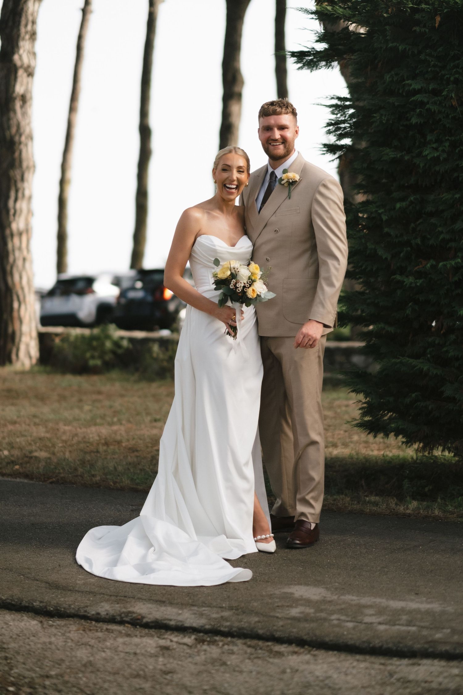 Smiling bride and groom posing together outdoors, bride in white strapless gown holding yellow bouquet.