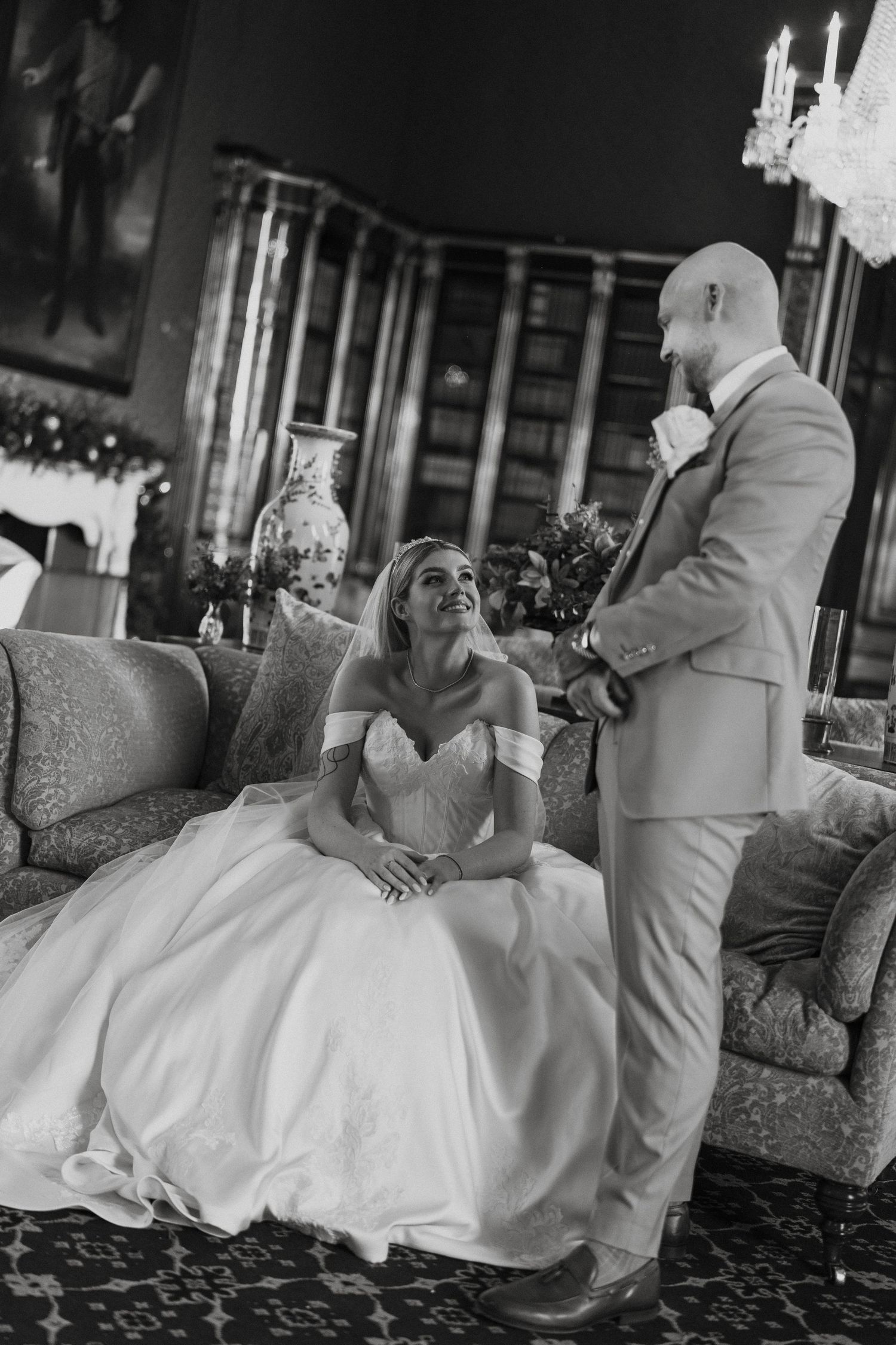 Black and white wedding photo of bride in ball gown seated on sofa smiling up at bald groom in suit in elegant room.