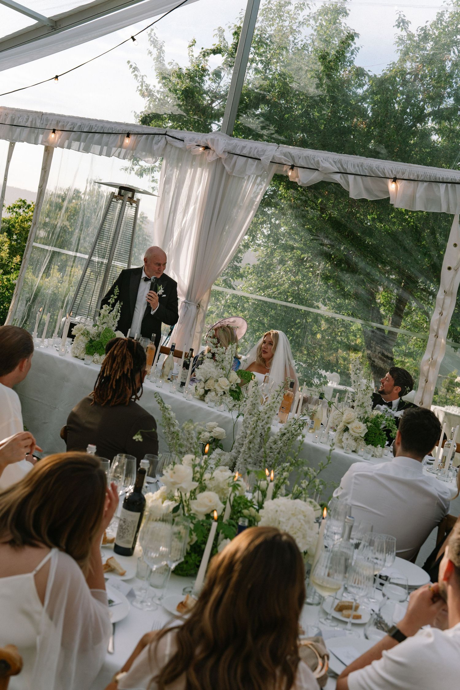 Wedding reception speech inside elegant clear tent with white floral arrangements and candlelit tables full of guests.